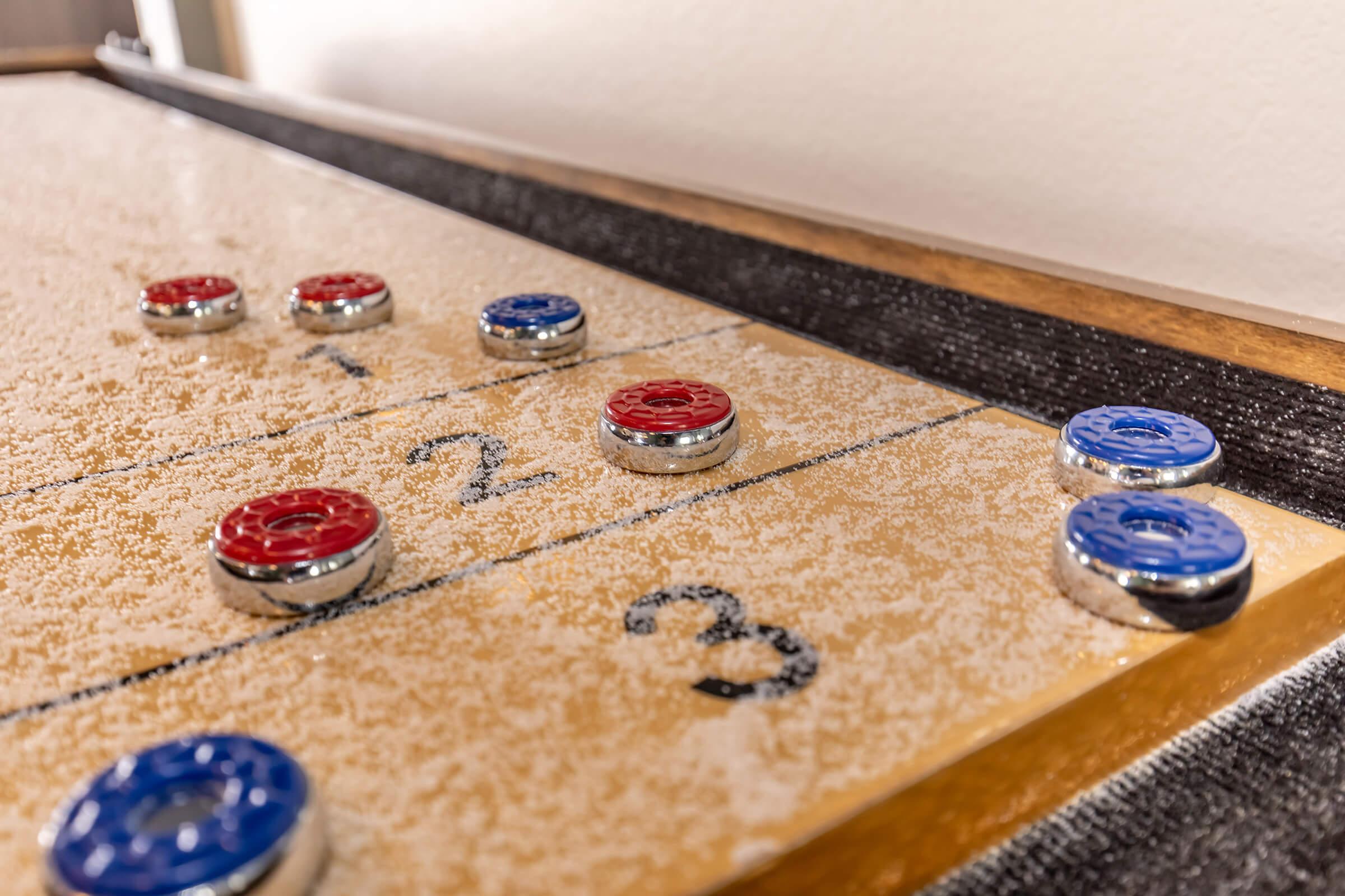 A close-up view of a shuffleboard table featuring red and blue pucks positioned on the playing surface. The table has scoring lines marked with numbers, and a fine layer of powder is visible on the surface, indicating recent gameplay. The background is softly blurred, emphasizing the table and pucks.