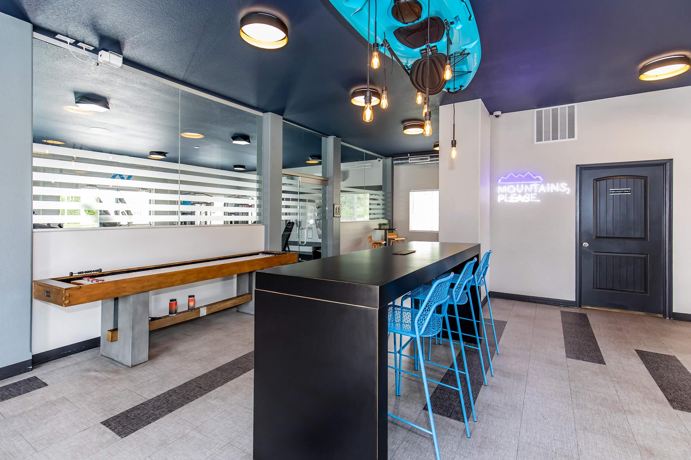 Modern reception area with a sleek black counter and blue bar stools. A wooden strip table is on the side, while frosted glass windows allow light in. Decorative lighting hangs from the ceiling, and a motivational sign on the wall reads "MOUNTAINS, PLEASE." The overall design features a contemporary aesthetic.