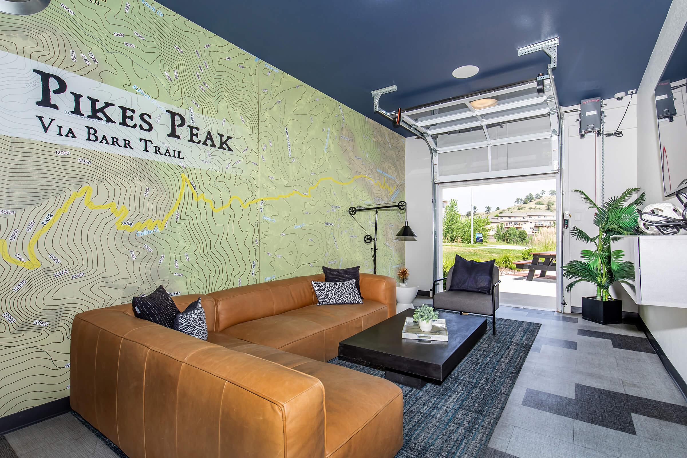 Stylish interior featuring a wall map of Pikes Peak, a brown leather sofa, a dark coffee table, and a cozy armchair. Natural light streams in from an open garage door, with greenery visible outside, creating a welcoming and modern atmosphere.