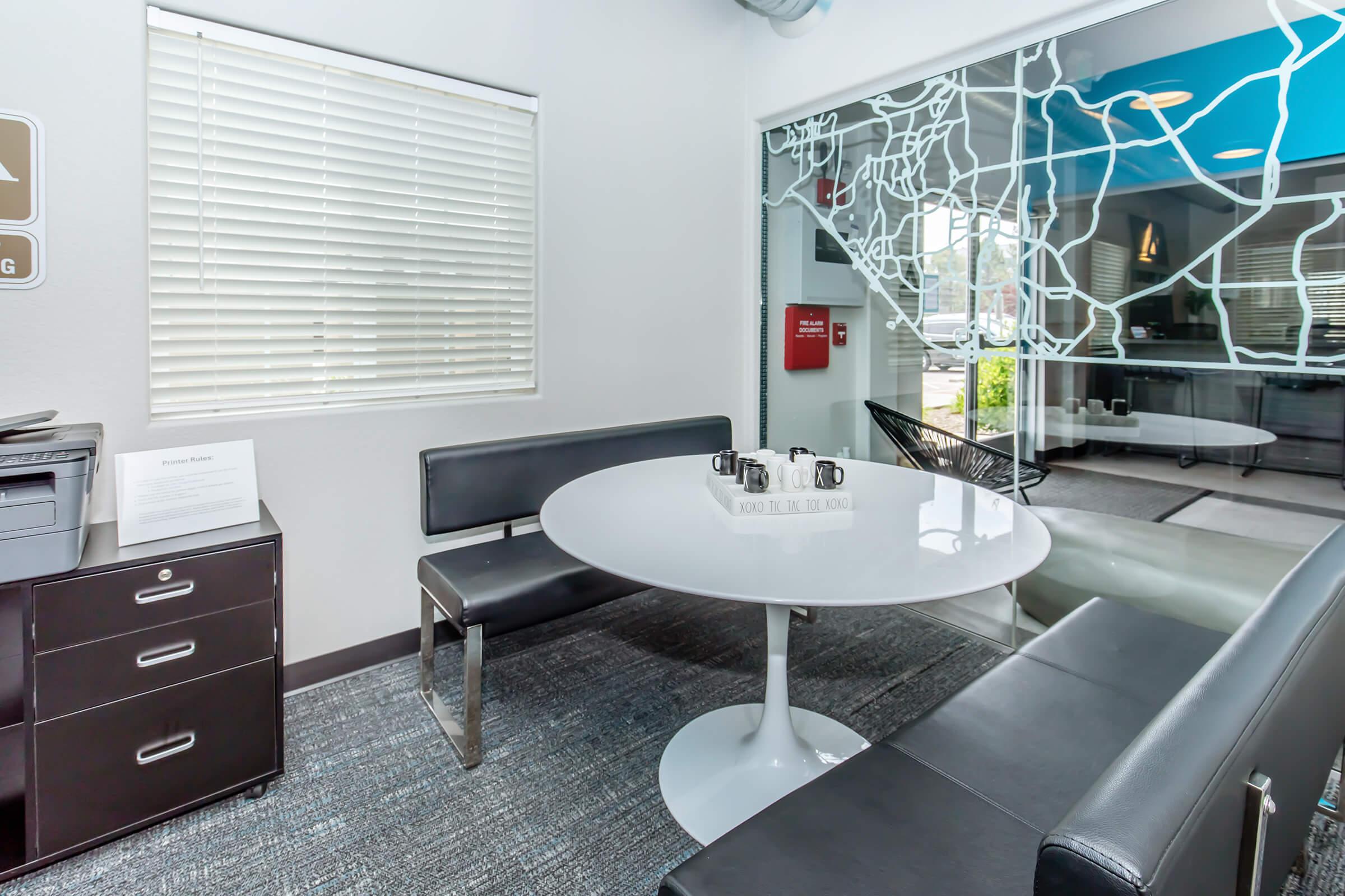 A modern office waiting area featuring a round white table with coffee cups, black leather seating, and a filing cabinet. The wall has an abstract map design, and a large window provides natural light, creating a welcoming atmosphere.