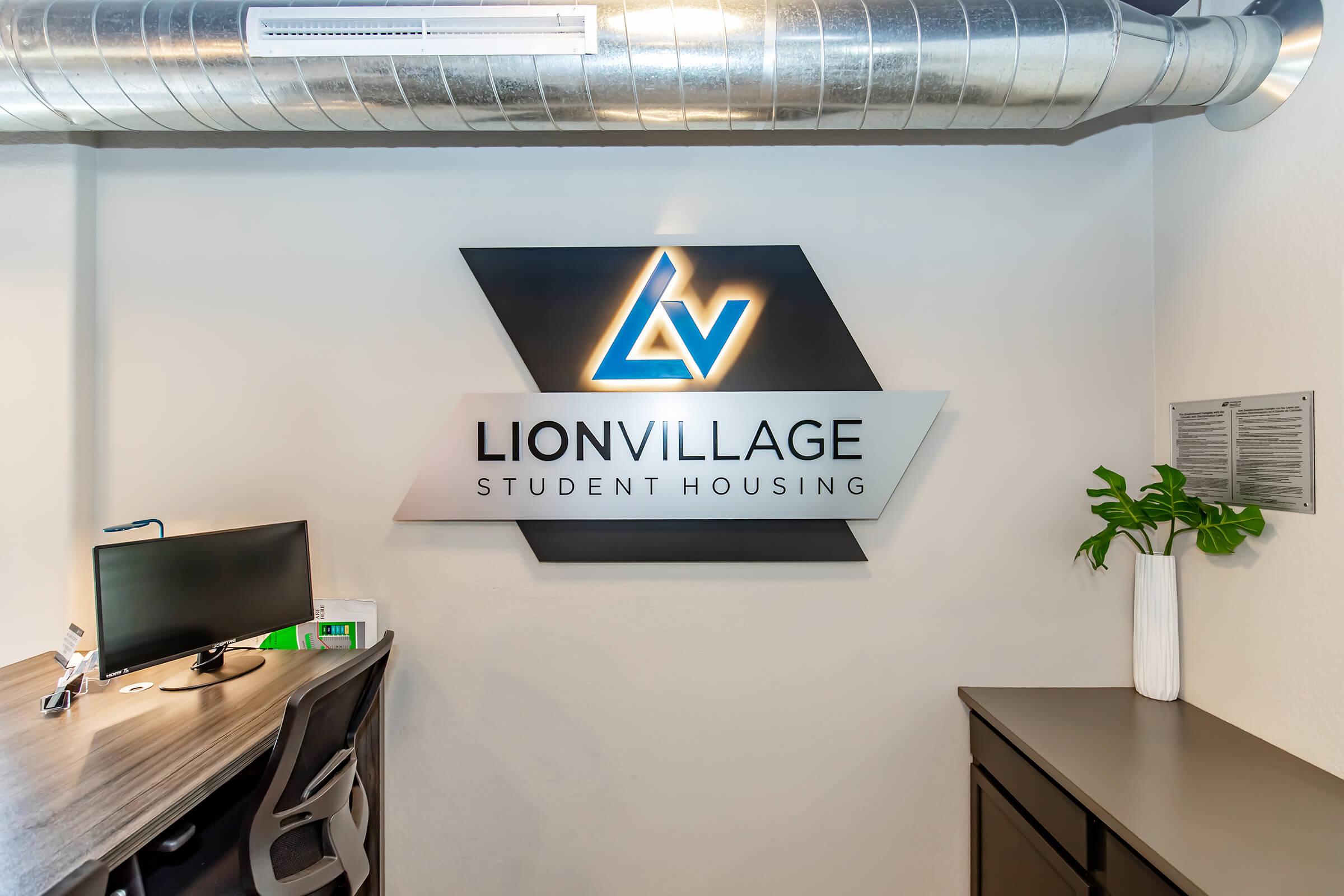 Signage for Lion Village Student Housing on a wall, featuring a sleek logo with a prominent blue and silver design. A desk with a computer is visible in the foreground alongside a plant in a modern white vase. The setting has industrial-style elements with visible metal ductwork above.