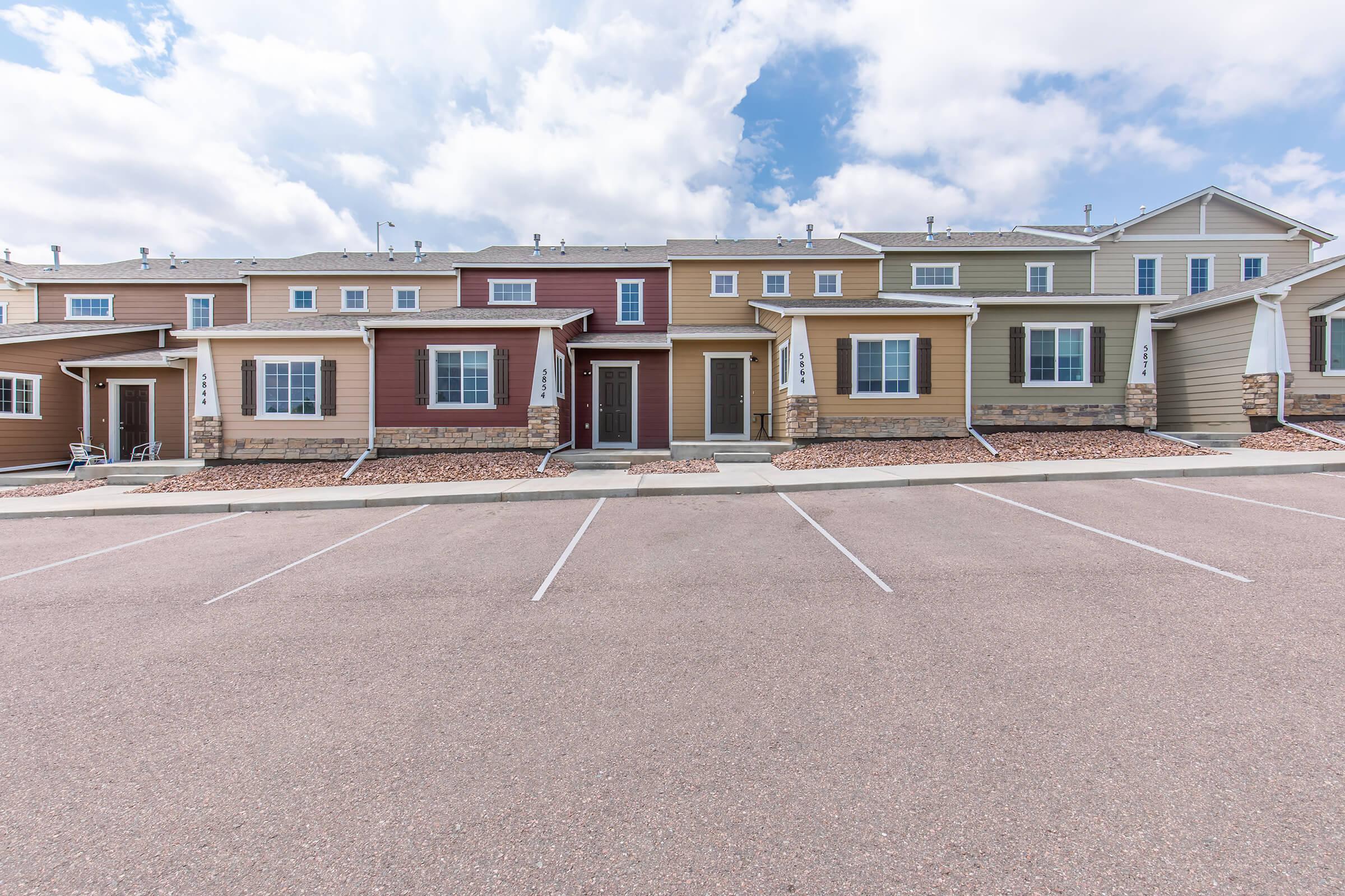 Row of multi-colored townhouse units with varying architectural styles, featuring front doors and windows. The buildings are set against a cloudy sky, and a paved parking area is visible in front of them. Decorative stone elements enhance the facades of some units.