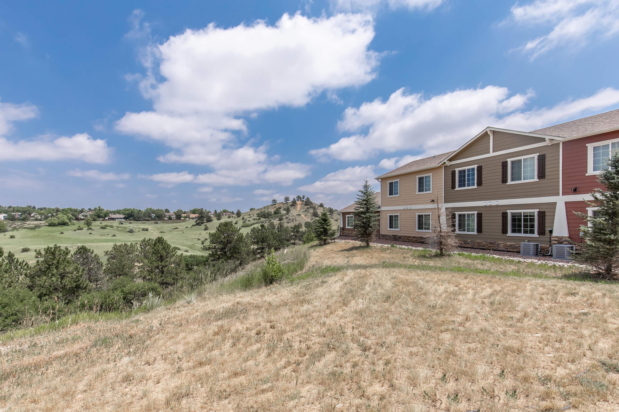 A view of a residential building with a mix of beige and pink siding, situated on a slight hill. The foreground features dry grass, while the background shows lush green trees and hills under a partly cloudy blue sky.