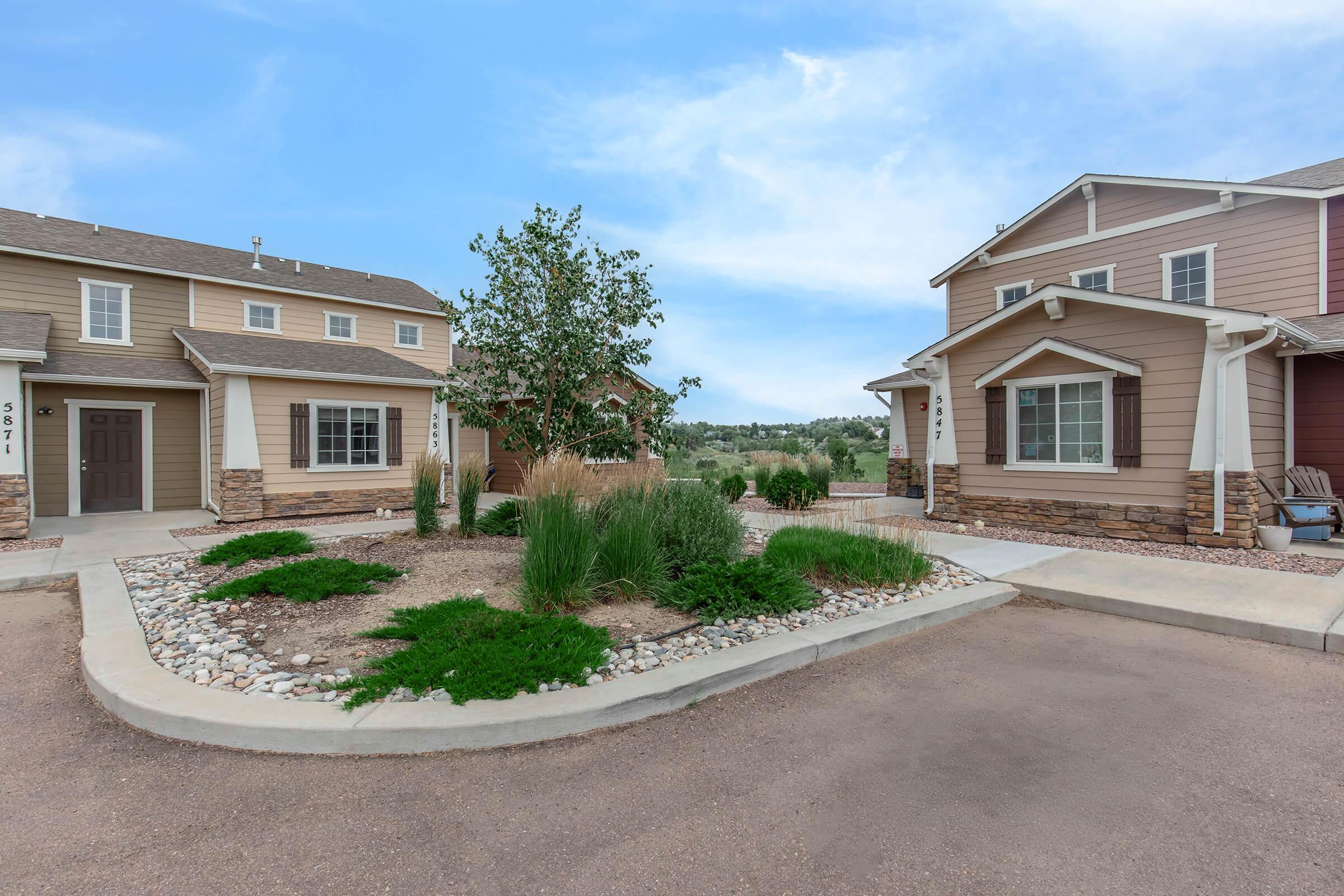 A landscaped residential area featuring two houses with different siding colors. The foreground includes a small garden with rocks and shrubs, while the background reveals green trees and hills. The scene is set under a clear blue sky, suggesting a pleasant day.