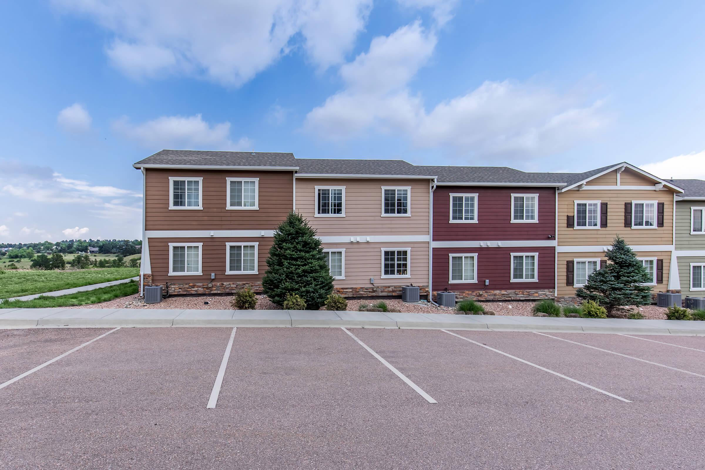 Row of three-story apartment buildings in a residential area, featuring a mix of beige, maroon, and light green siding. The foreground shows a parking lot with empty spaces, and the background includes a grassy area and a clear blue sky with scattered clouds.