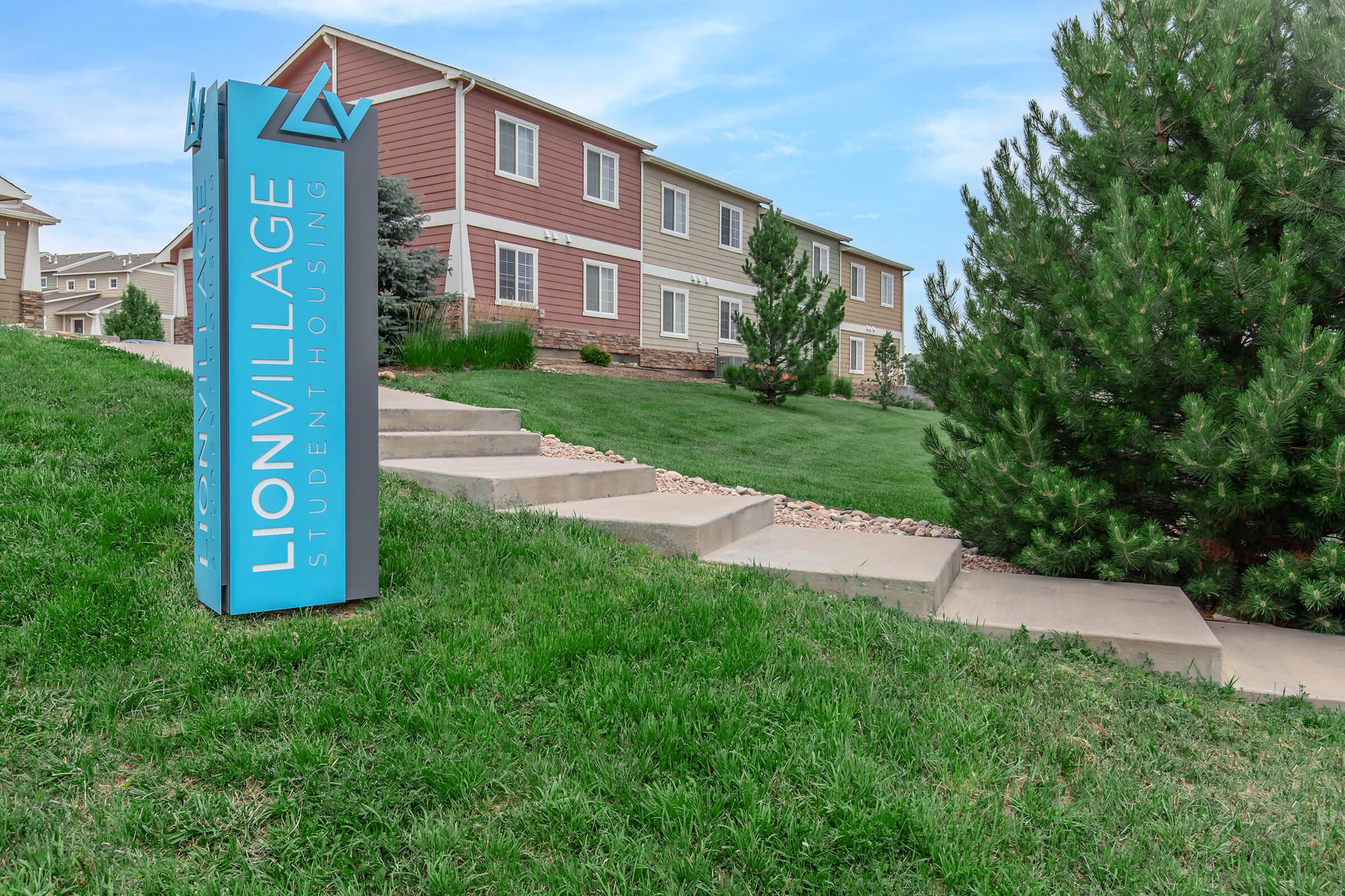 A sign for Lion Village Student Houses is prominently displayed in front of a well-maintained campus area. The background features a row of multi-colored residential buildings with green lawns and shrubs. The sky is clear, adding to the inviting atmosphere of the student housing complex.