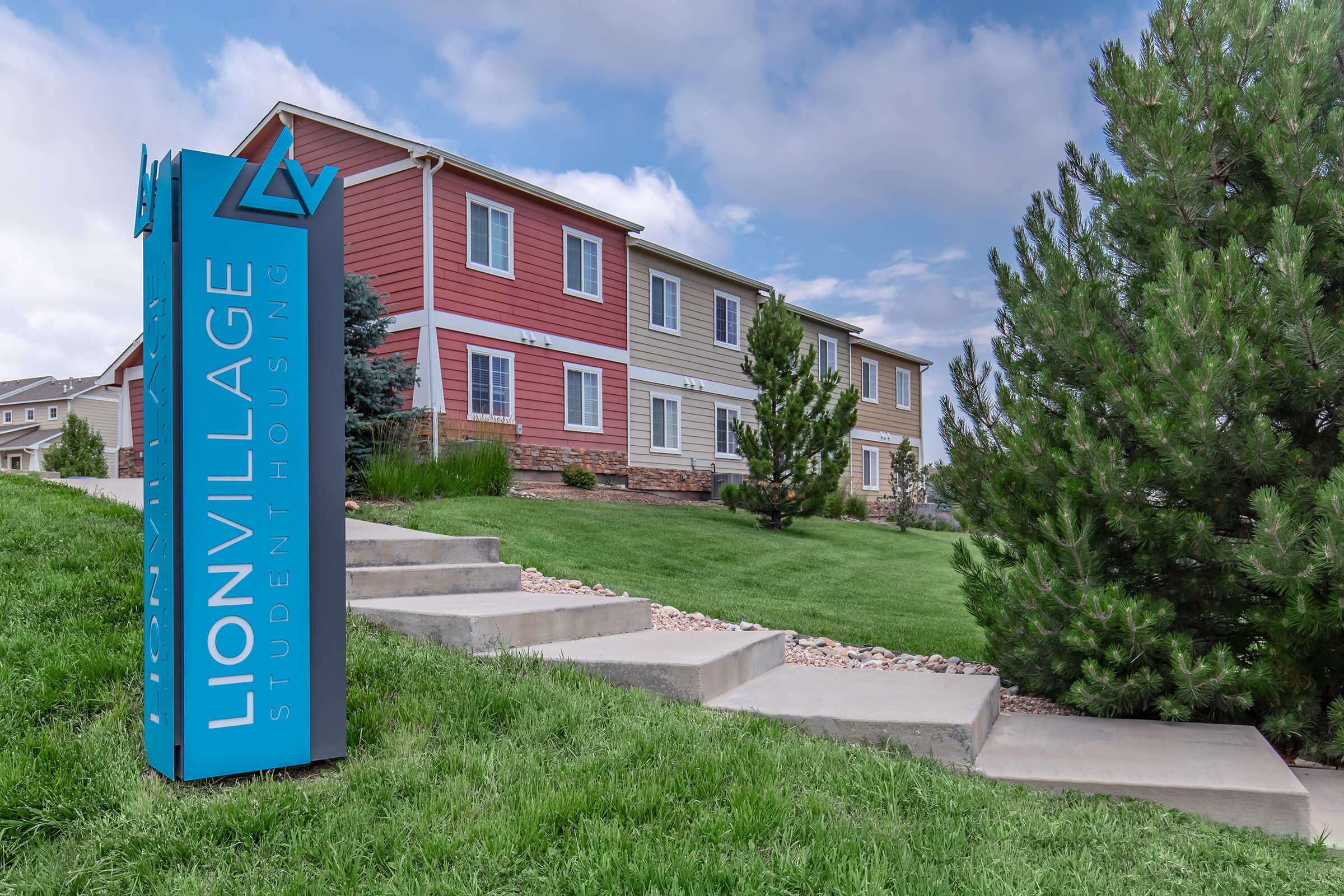 Sign for Lion Village Student Housing in front of a two-story building with multiple windows. The building features a mix of red, beige, and white siding, surrounded by green grass and trees. The sky is partly cloudy.