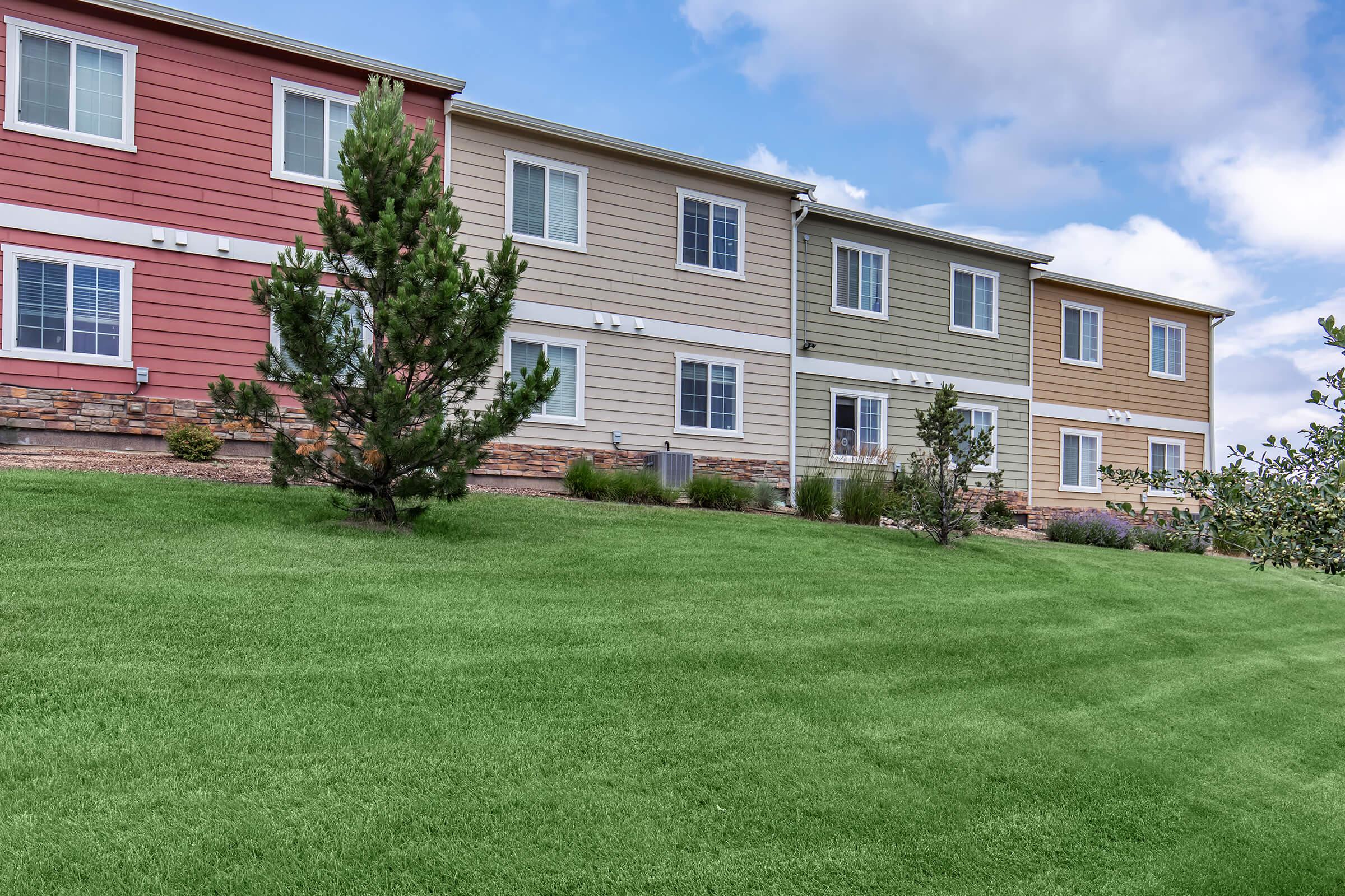 Row of multi-colored apartment buildings with a green lawn in the foreground and a blue sky with clouds in the background. The buildings feature various siding colors, including red, beige, and green, with windows and landscaping that adds a neat appearance.