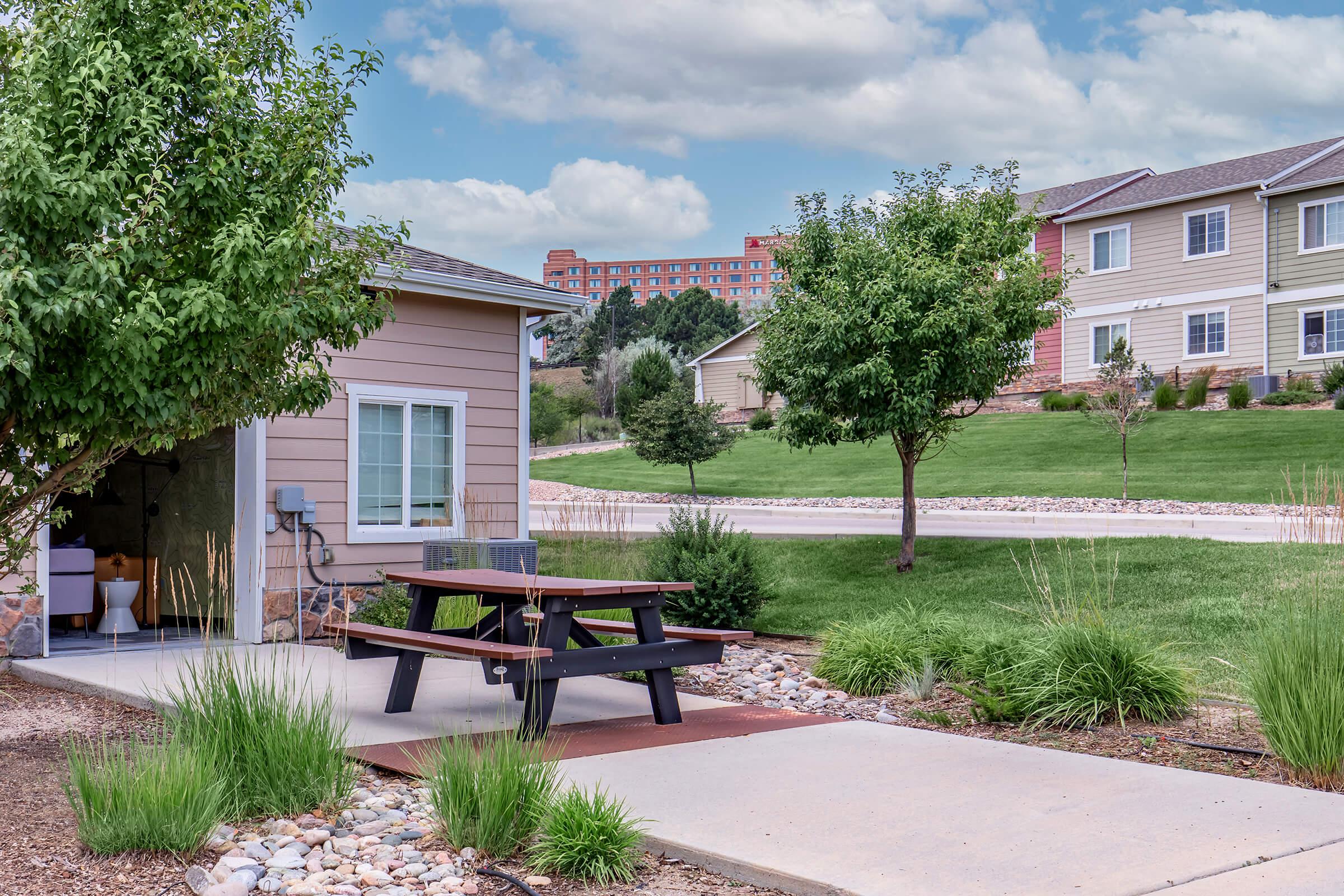 A well-maintained outdoor area featuring a picnic table on a concrete pad surrounded by grass and landscaped greenery. In the background, there are residential buildings and a distant hotel against a partly cloudy sky.