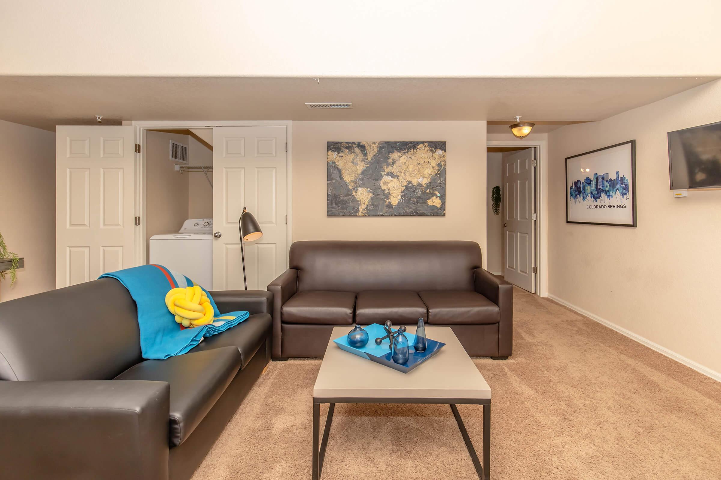 A cozy living area featuring a dark leather sofa, a second sofa with a colorful throw, and a coffee table with decorative items. The walls are adorned with a world map and framed artwork. In the background, there are closed doors and a laundry area, contributing to a homey atmosphere. Soft lighting enhances the space.