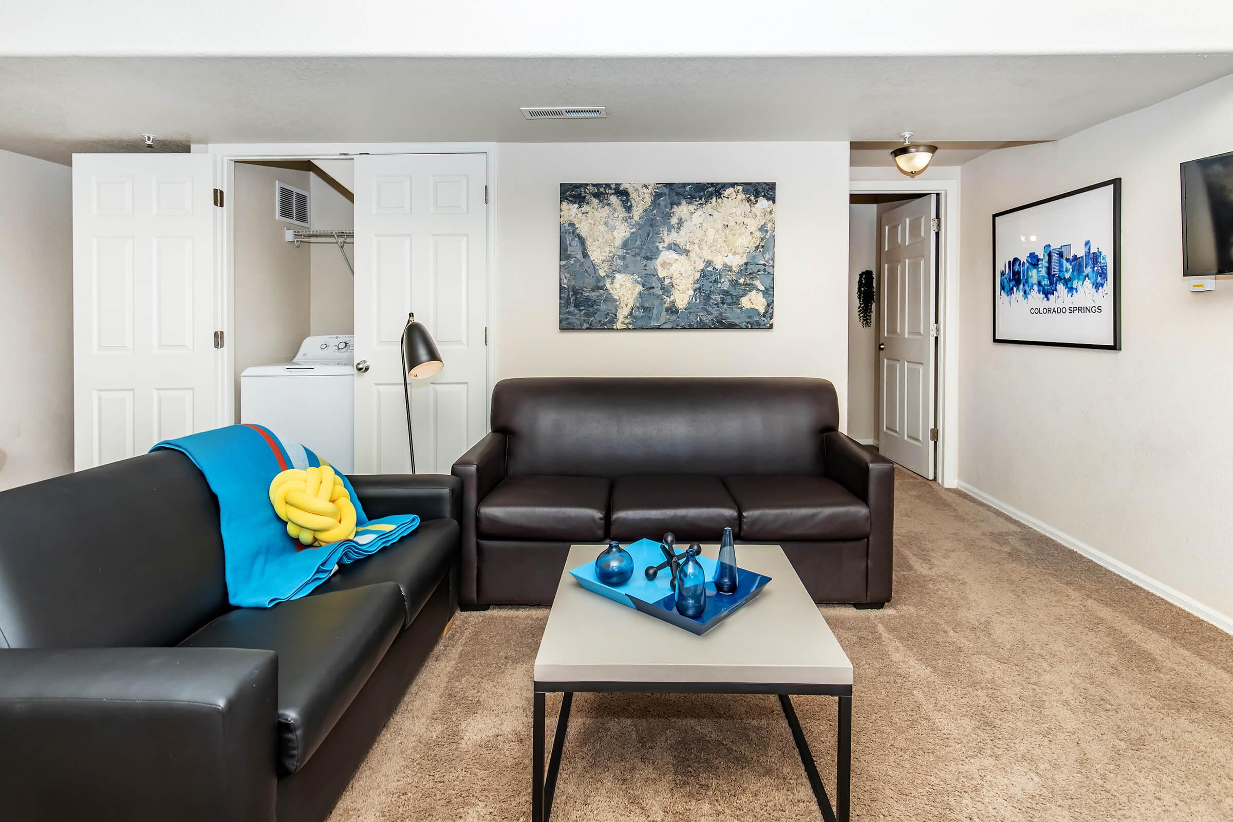 A cozy living room featuring a dark leather sofa, a contrasting black coffee table with decorative items, and a vibrant blue throw on the couch. The room includes a framed world map on the wall, light carpeting, and access to a laundry area through double doors. Natural light fills the space, creating a welcoming atmosphere.