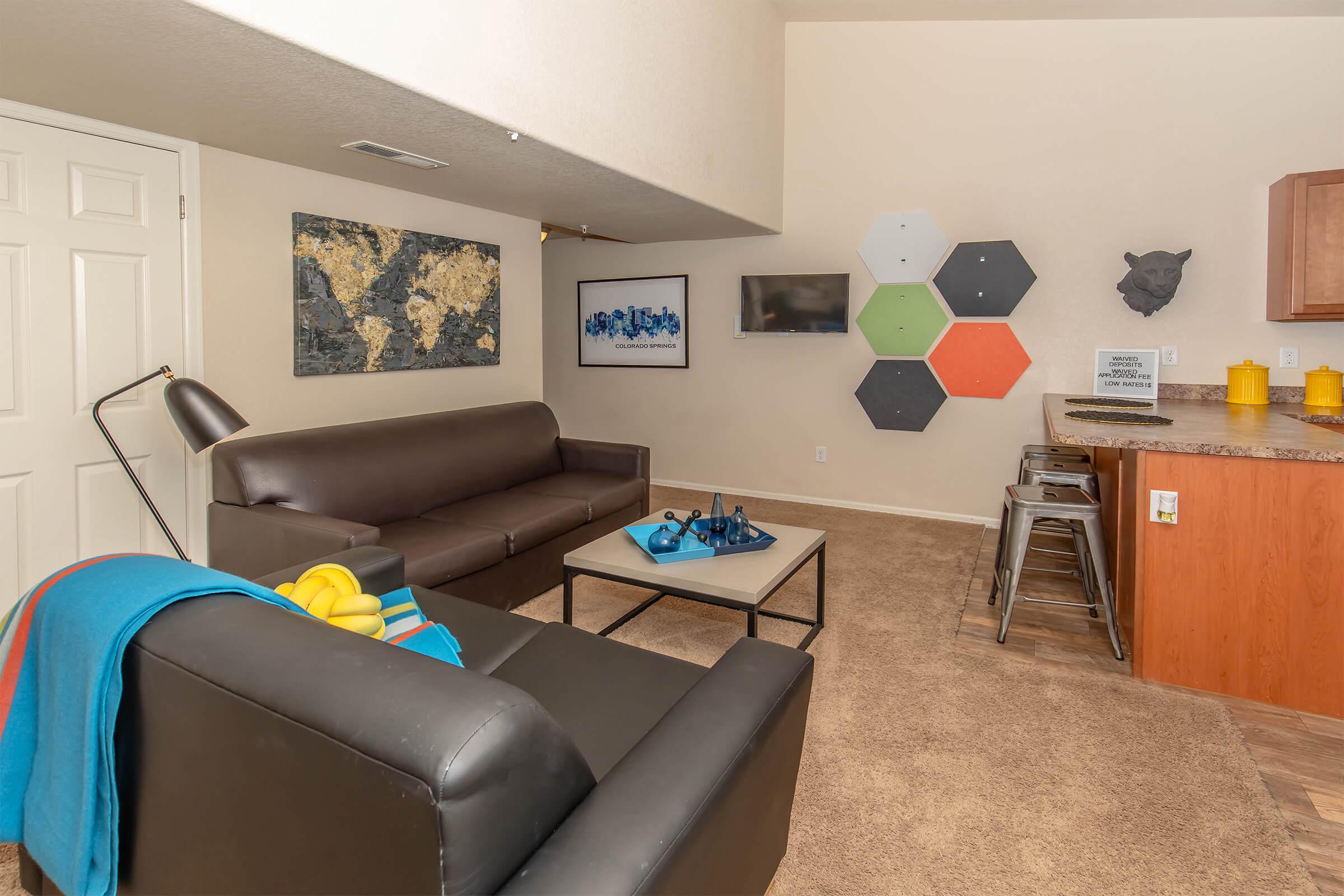 A modern living room featuring two black sofas, a coffee table, and a colorful wall decor. One wall displays a world map and another features hexagonal shapes in various colors. There's a flat-screen TV mounted on the wall and a small bar area with stools. The carpeted floor adds a cozy touch to the space.
