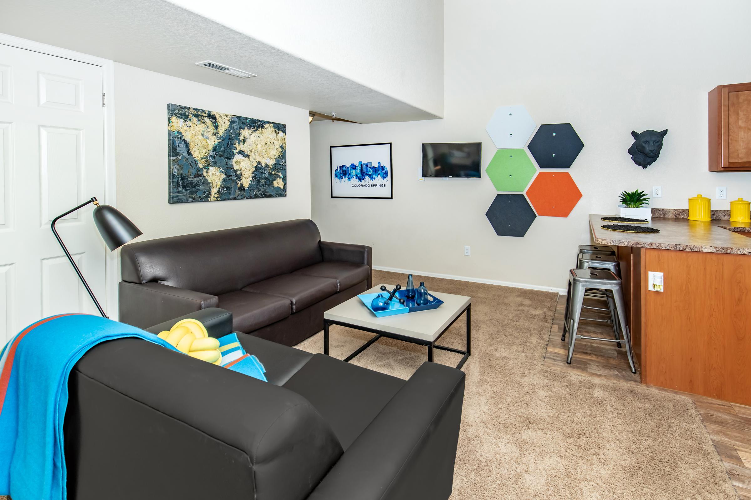 A modern living room featuring a black leather sofa with a colorful throw blanket, a coffee table, and a kitchenette. The walls are adorned with abstract art, a wall-mounted TV, and decorative hexagonal pieces. A cozy, inviting atmosphere with neutral carpeting and light-colored walls.