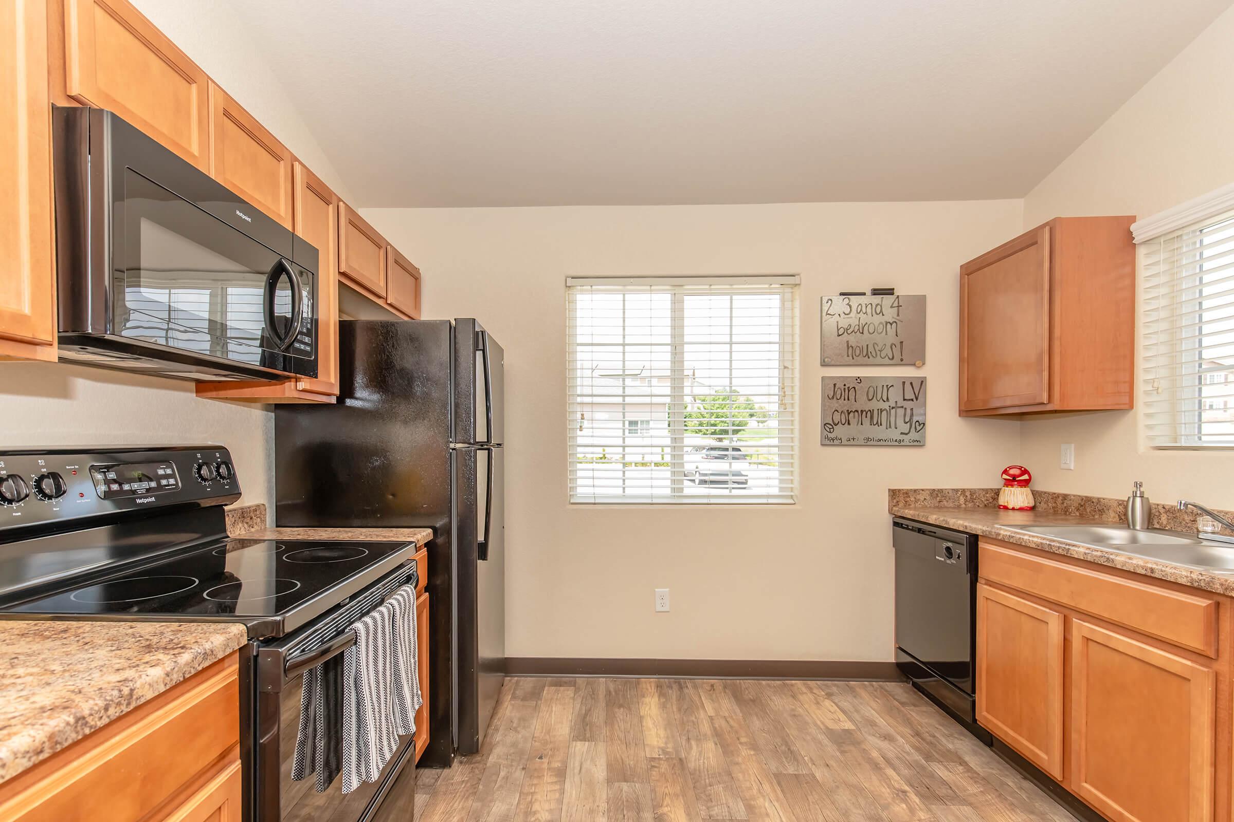 Modern kitchen with wood cabinetry, black appliances including a microwave, stove, and refrigerator. Countertops are a light brown speckled design. A window provides natural light, and there is a decorative sign on the wall with text. The flooring is a wood-like laminate.