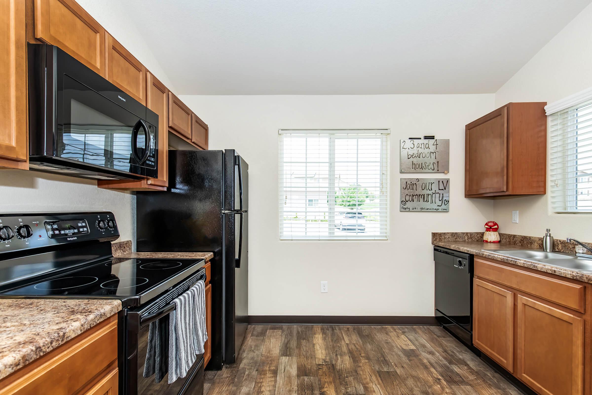 A modern kitchen featuring wooden cabinets, a granite countertop, and stainless steel appliances. The black refrigerator, oven, and microwave are neatly arranged. A window allows natural light, and a decorative sign on the wall invites viewers to "Join our community." The floor is a dark wood laminate.