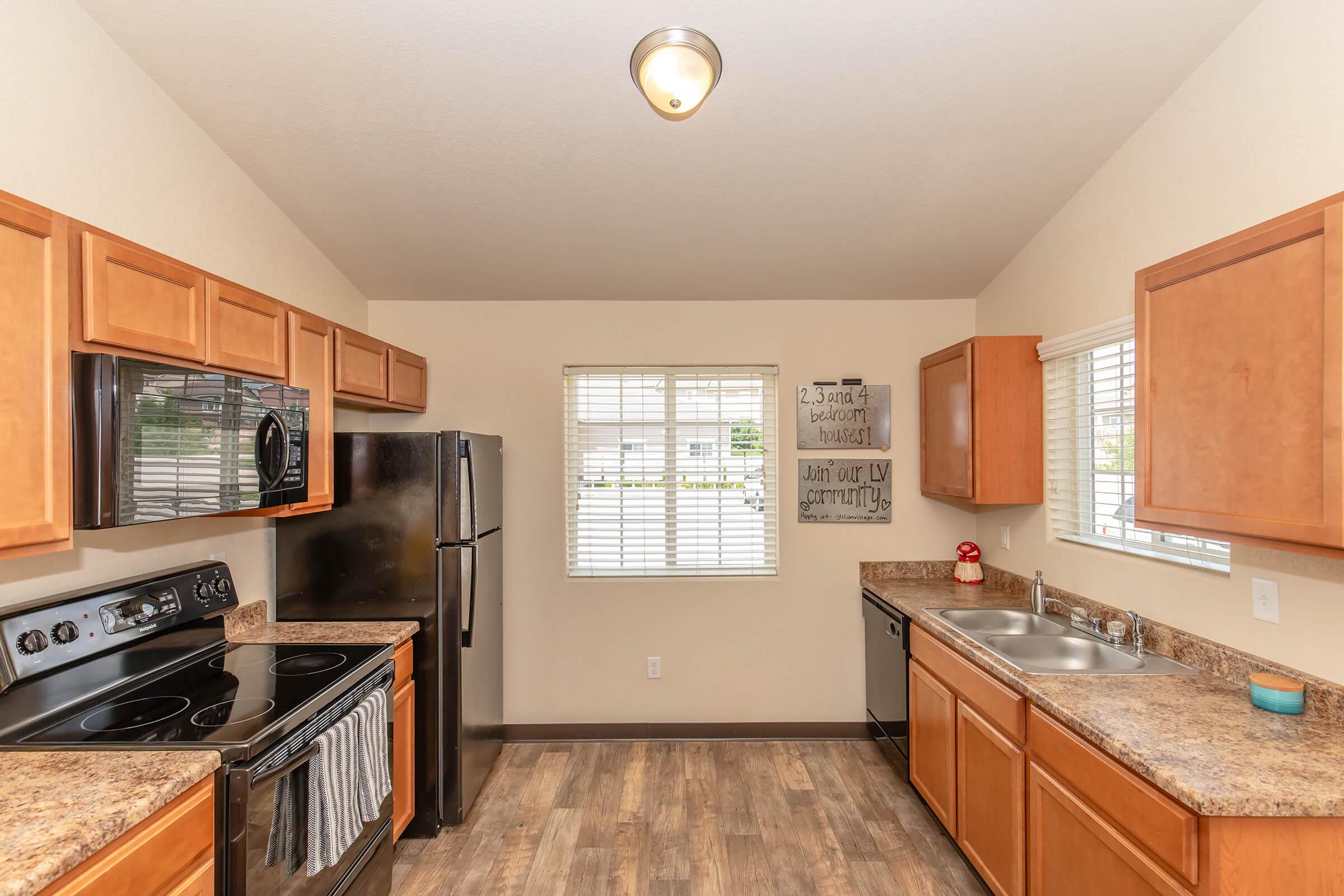 Kitchen with wooden cabinets, black appliances including an oven, stove, and refrigerator. A double sink is under a window with blinds. There are light-colored granite countertops and a decorative wall sign. The floor has a wood-like finish, and the lighting is bright from a ceiling fixture.