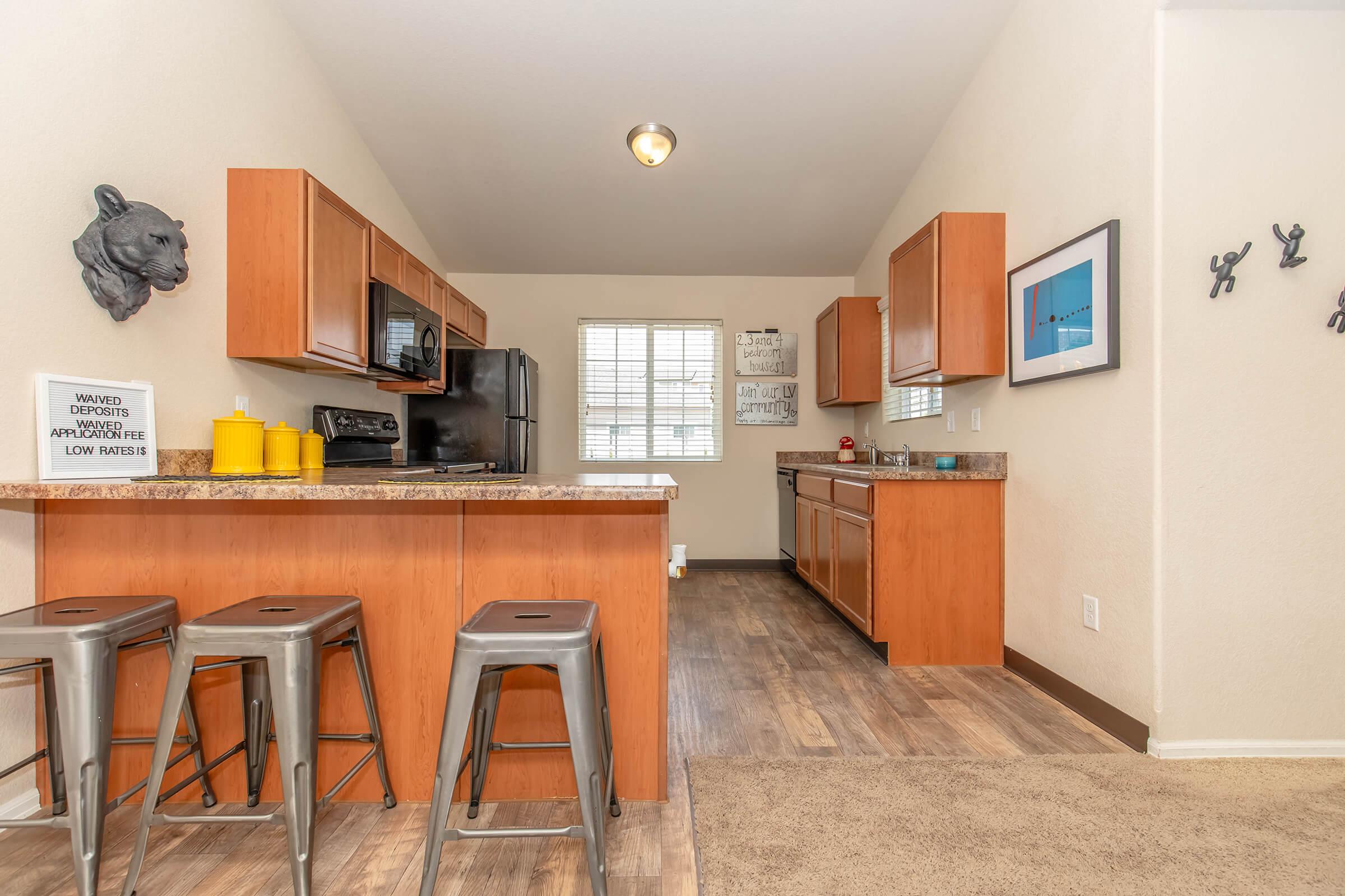 A modern kitchen featuring wooden cabinets, a granite countertop, and stainless steel appliances. Three metal bar stools are arranged around the counter. The space is well-lit with natural light from a window, and there is a decorative wall piece of a lion's head along with framed art on the walls.