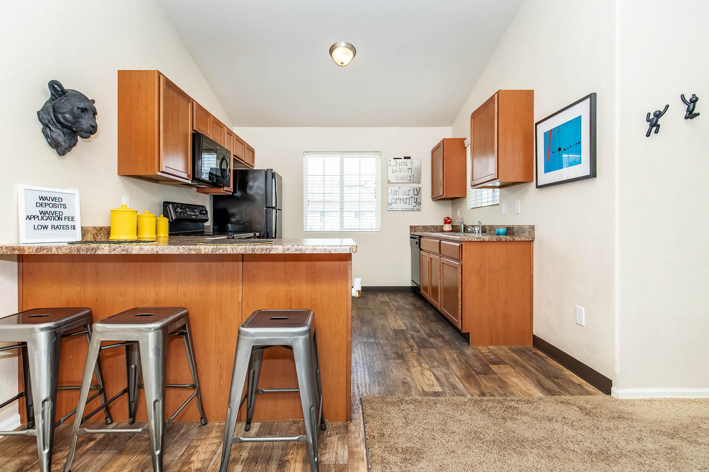 A modern kitchen featuring wooden cabinets, stainless steel appliances, and a small breakfast bar with metallic stools. A window allows natural light, and decorative elements include wall art and a small sign. The flooring is a mix of wood and carpet, creating a cozy atmosphere.