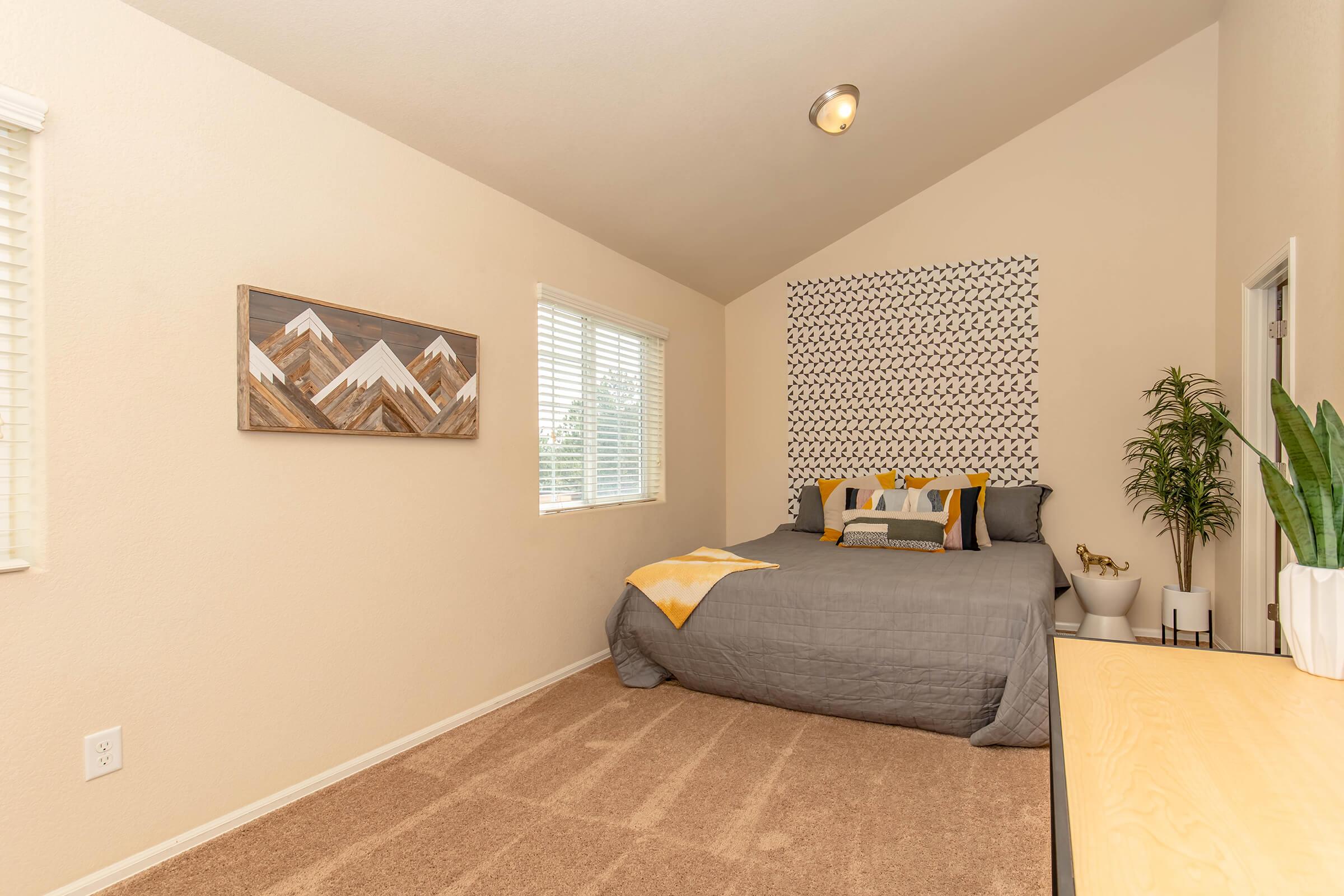A cozy bedroom featuring a gray bedspread with decorative pillows, a geometric accent wall with mountain patterns, a potted plant, and a wooden dresser. Large windows allow natural light to fill the space, highlighting the neutral-colored walls and soft carpet flooring.