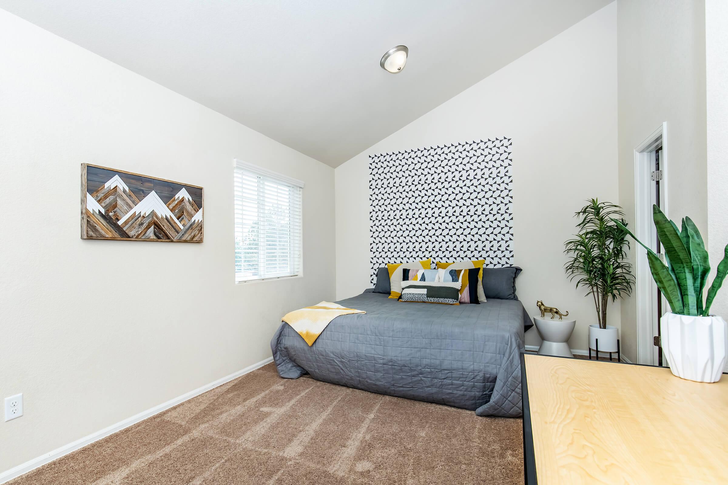 A cozy bedroom featuring a queen-sized bed with gray bedding and decorative pillows. The walls are painted light colors, and a geometric patterned accent wall adds charm. A window lets in natural light, and there's a potted plant in the corner. A wooden dresser is visible, enhancing the room's modern aesthetic.