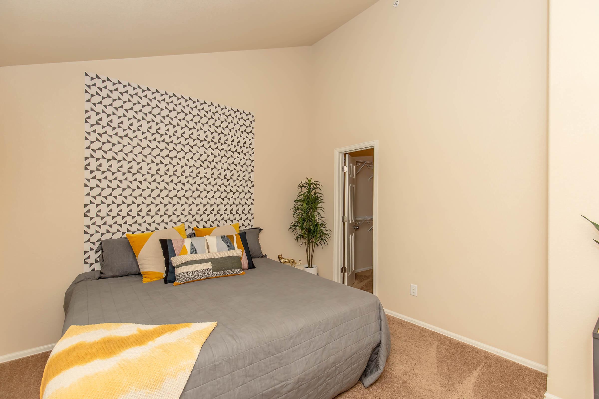 A cozy bedroom featuring a neatly made bed with gray bedding and decorative pillows in yellow and white. The wall behind the bed has a geometric patterned design. A potted plant is positioned beside the bed, and a doorway leads to another room. The walls are a soft beige color, enhancing the warm ambiance.