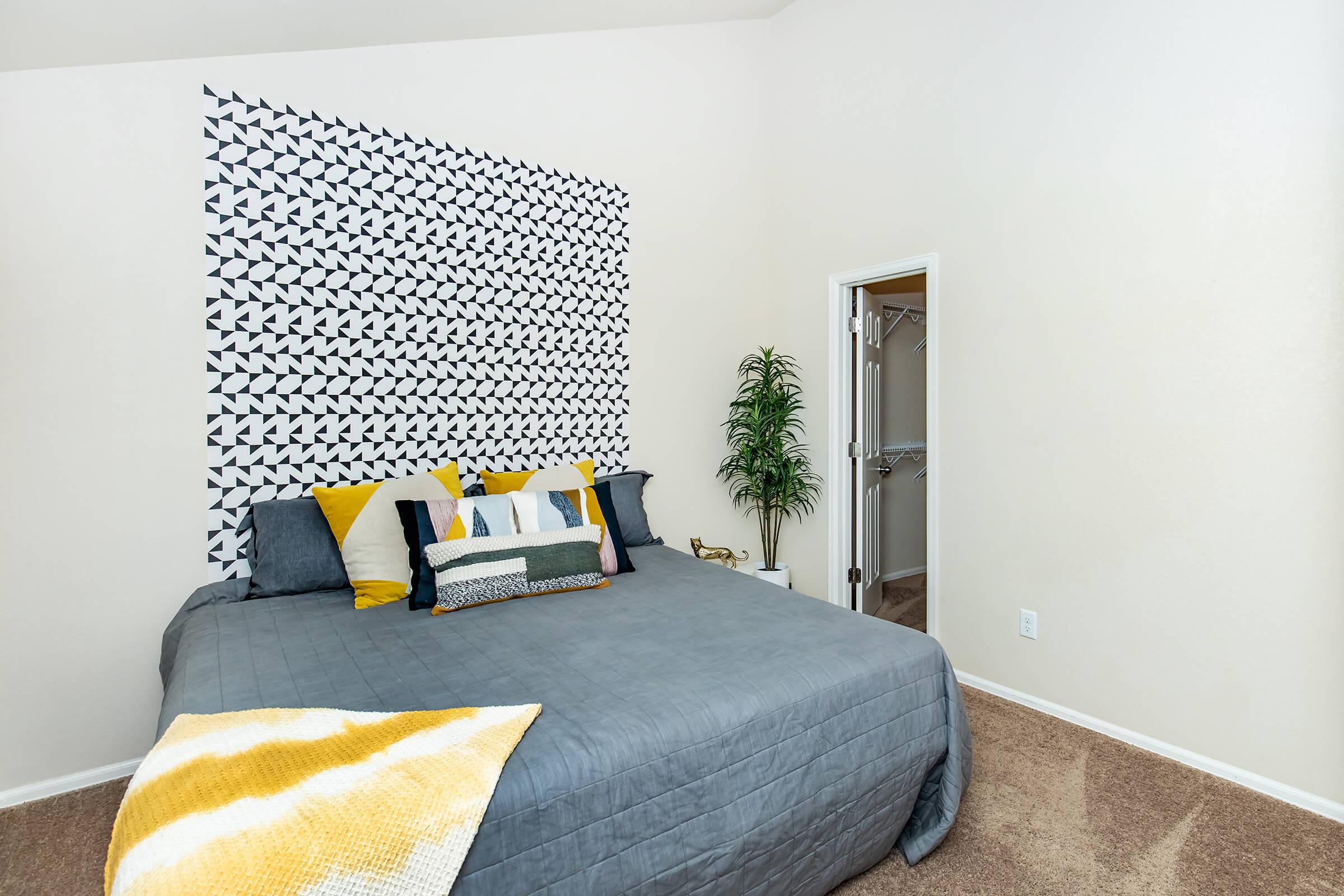 A cozy bedroom featuring a large bed with gray bedding and colorful decorative pillows. The wall behind the bed is adorned with a geometric black and white pattern. A potted plant adds a touch of greenery, while a partially open door reveals a closet space. The room has neutral-colored walls and soft carpet flooring.