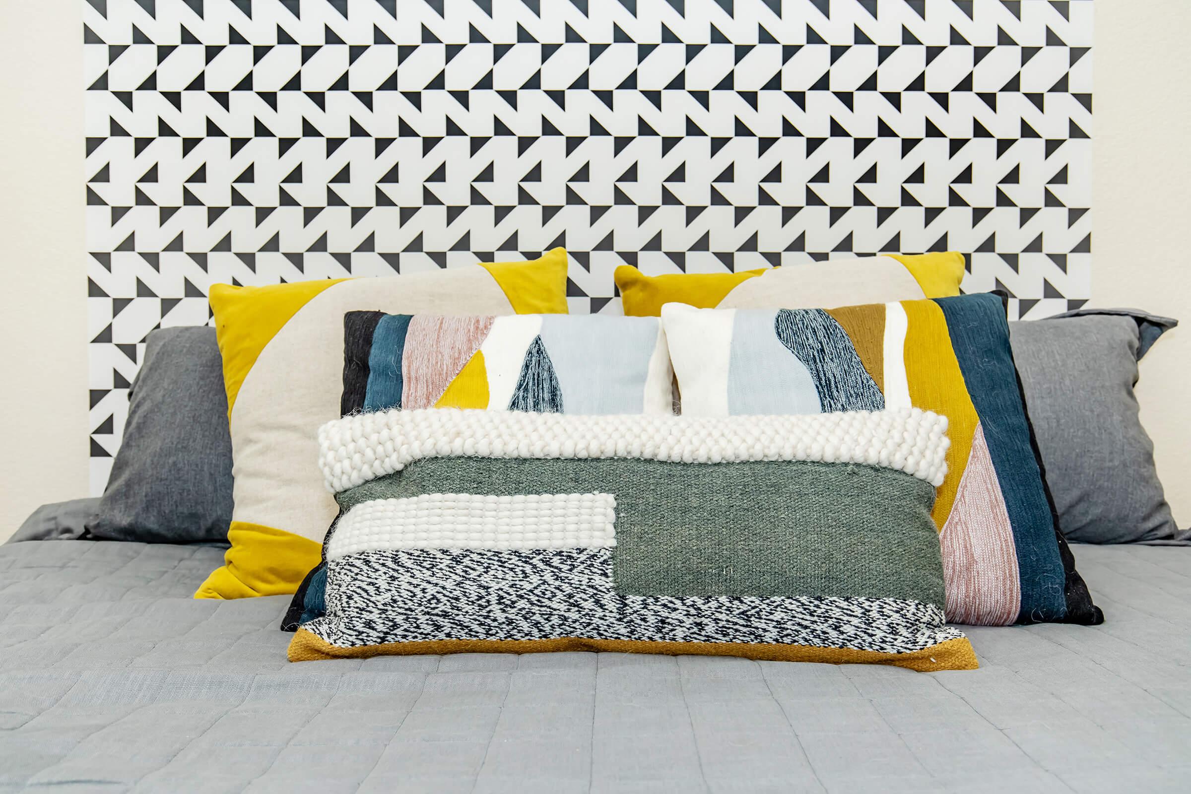 A neatly made bed featuring a variety of decorative pillows in earthy tones and patterns. The headboard is adorned with a geometric black and white design, complementing the colorful pillows arranged in a layered fashion on a textured gray bedspread.