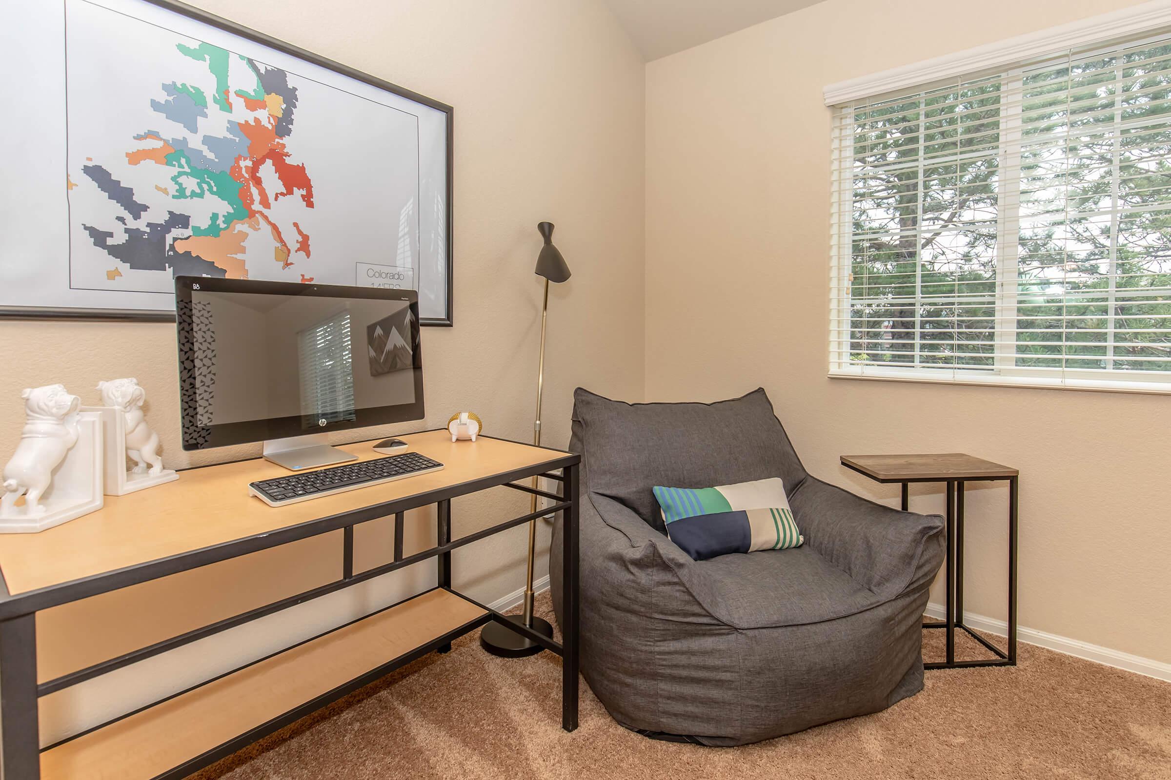 Home office setup featuring a modern desk with a computer monitor, a cozy bean bag chair with a striped pillow, and a small side table. The wall displays a colorful abstract map print, and a window allows natural light to enter the room, enhancing the warm atmosphere.