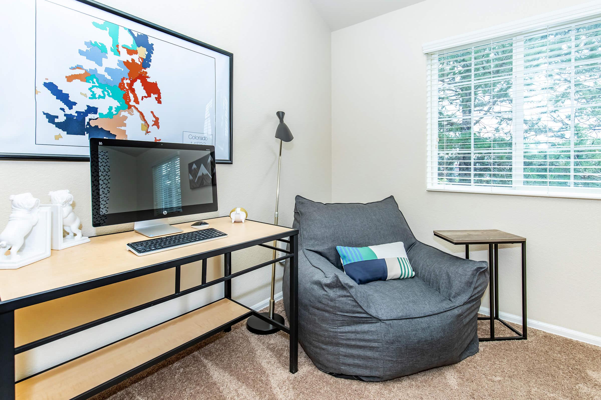 A cozy home office featuring a modern desk with a computer, a bean bag chair, and a small side table. A colorful abstract map is framed on the wall, and natural light streams in through a window adorned with blinds. The carpet is a neutral tone, creating a warm ambiance.