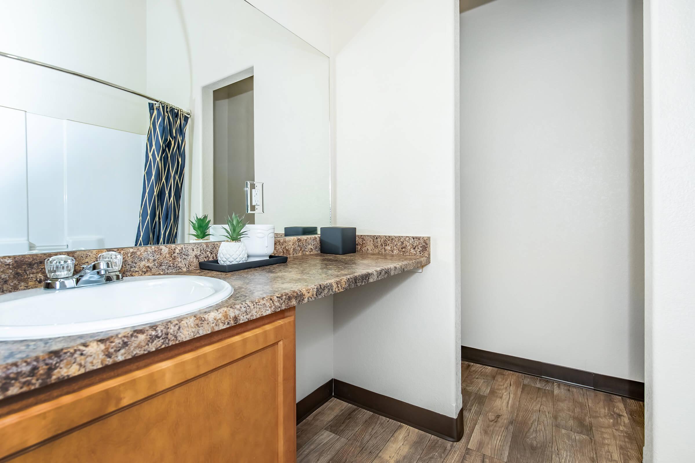 A clean and modern bathroom with a wooden vanity featuring a white sink. There are decorative plants on the countertop, a shower curtain with a blue and gold pattern, and a light-colored wall. The floor is made of wooden planks, and a door leads to another space.
