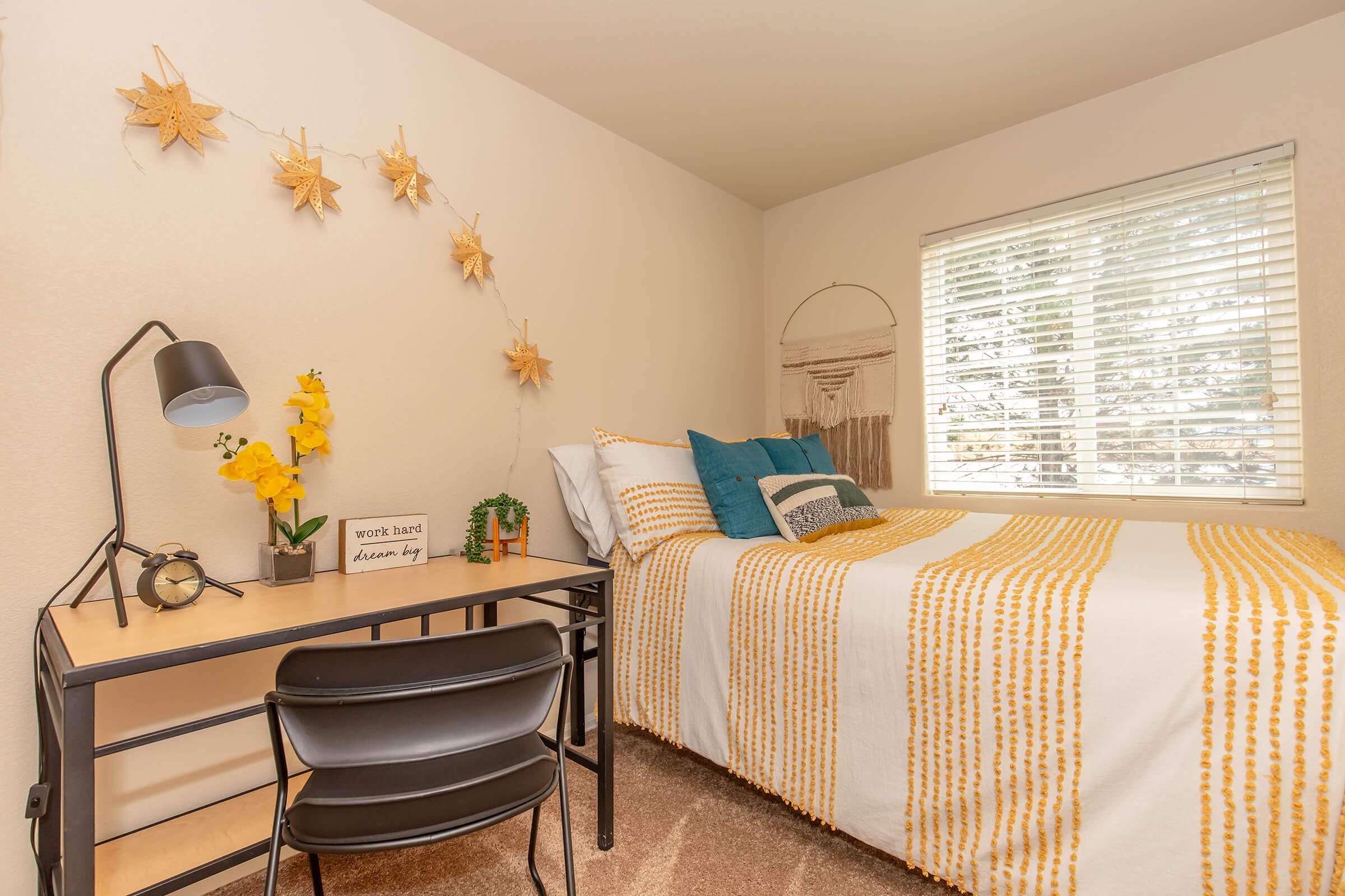 A cozy bedroom with a bed dressed in yellow and white striped bedding. There's a desk with a lamp and a small plant, along with decorative elements like a sign that says "work hard dream big" and hanging star-shaped ornaments on the wall. A window lets in natural light, enhancing the inviting atmosphere.