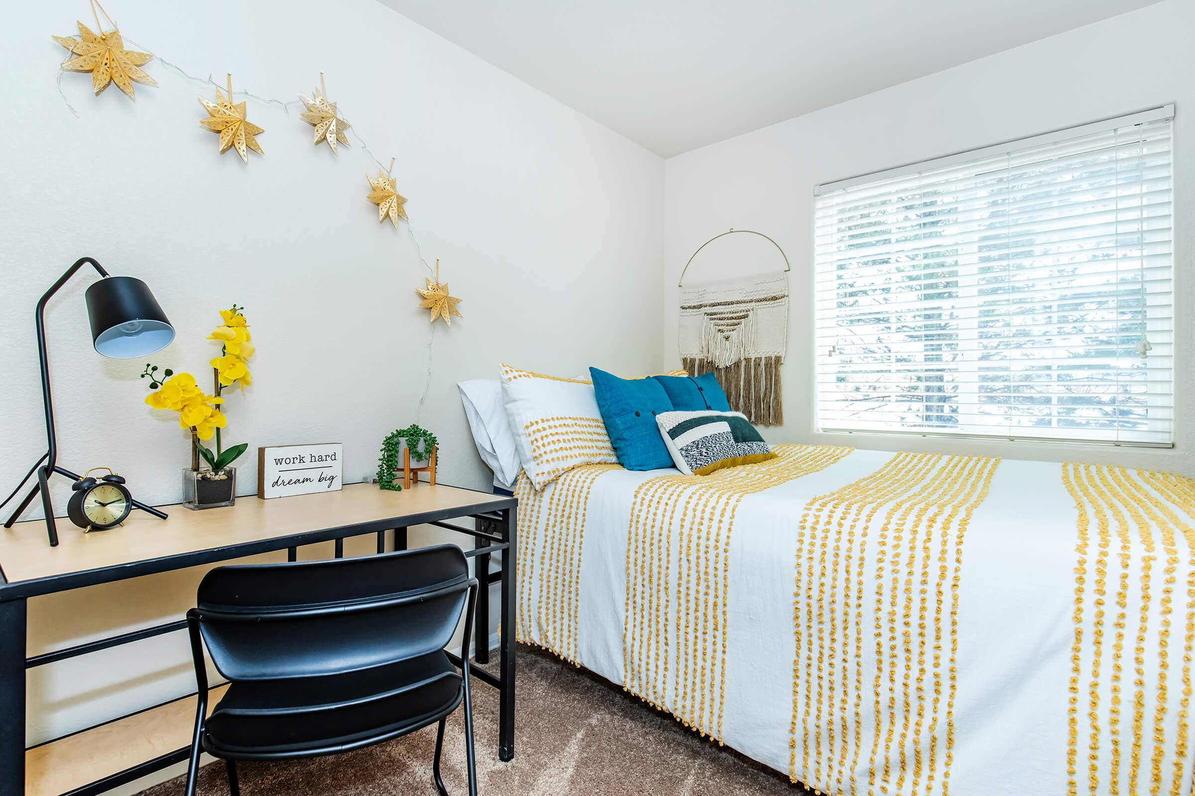 A cozy bedroom featuring a neatly made bed with a yellow and white striped comforter, accented by decorative pillows. A desk with a lamp and a small plant is positioned nearby. The wall is adorned with star decorations, and sunlight filters through a window covered with blinds. A motivational sign sits on the desk.
