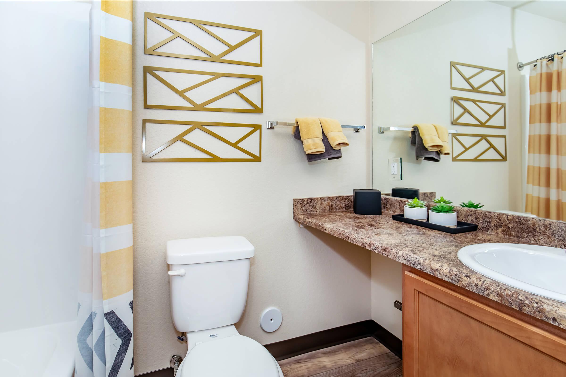 A modern bathroom featuring a white toilet, a granite countertop with a sink, and a mirror. Yellow towels hang on a wall rack, and decorative succulents are placed on the counter. The shower curtain displays a geometric pattern, and the walls are painted light beige.