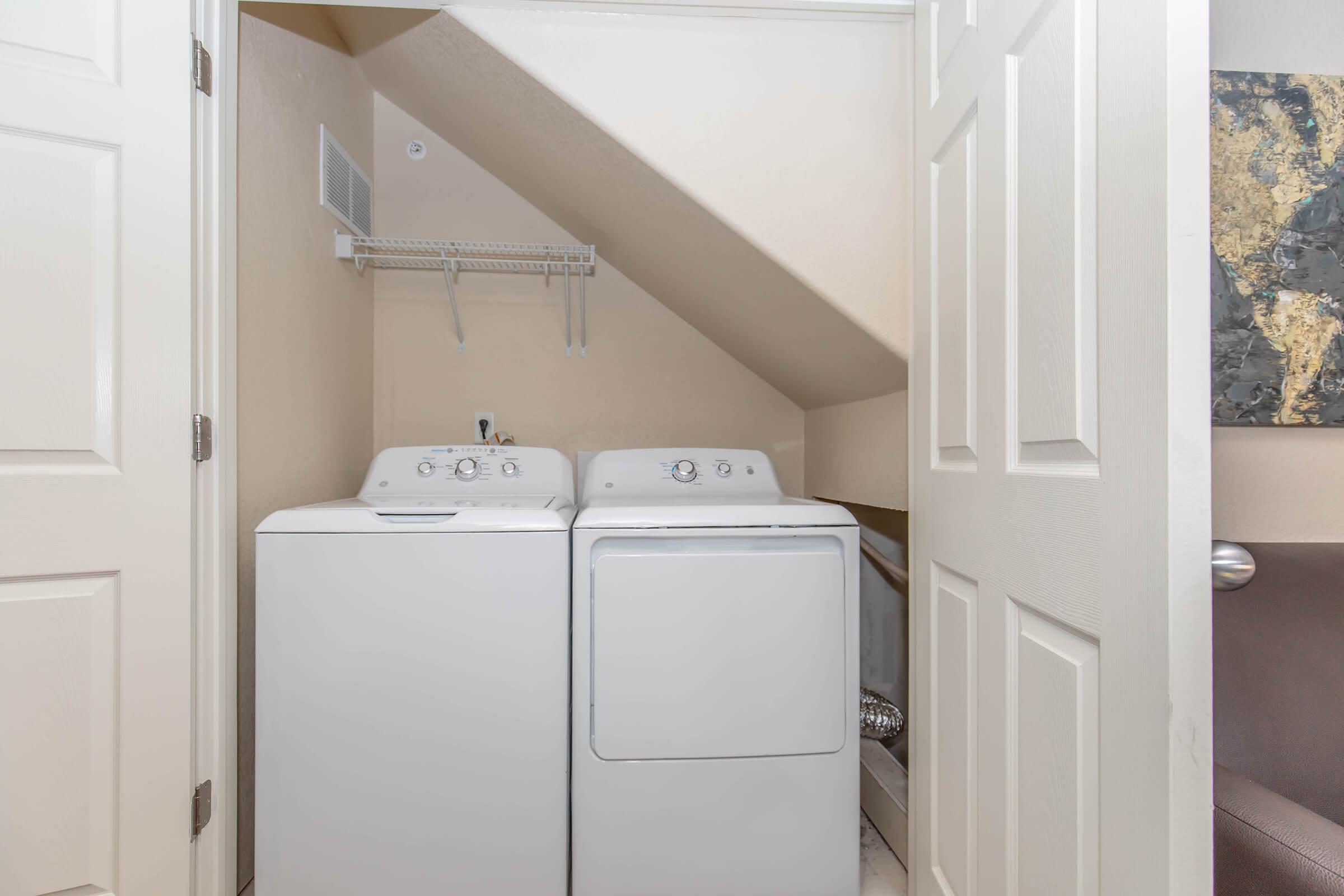 A laundry room tucked under a staircase features a white washer and dryer side by side. The walls are a light beige color, and there is a hanging shelf above the appliances. A door partially opens to an adjacent room, adding to the compact space's functionality.