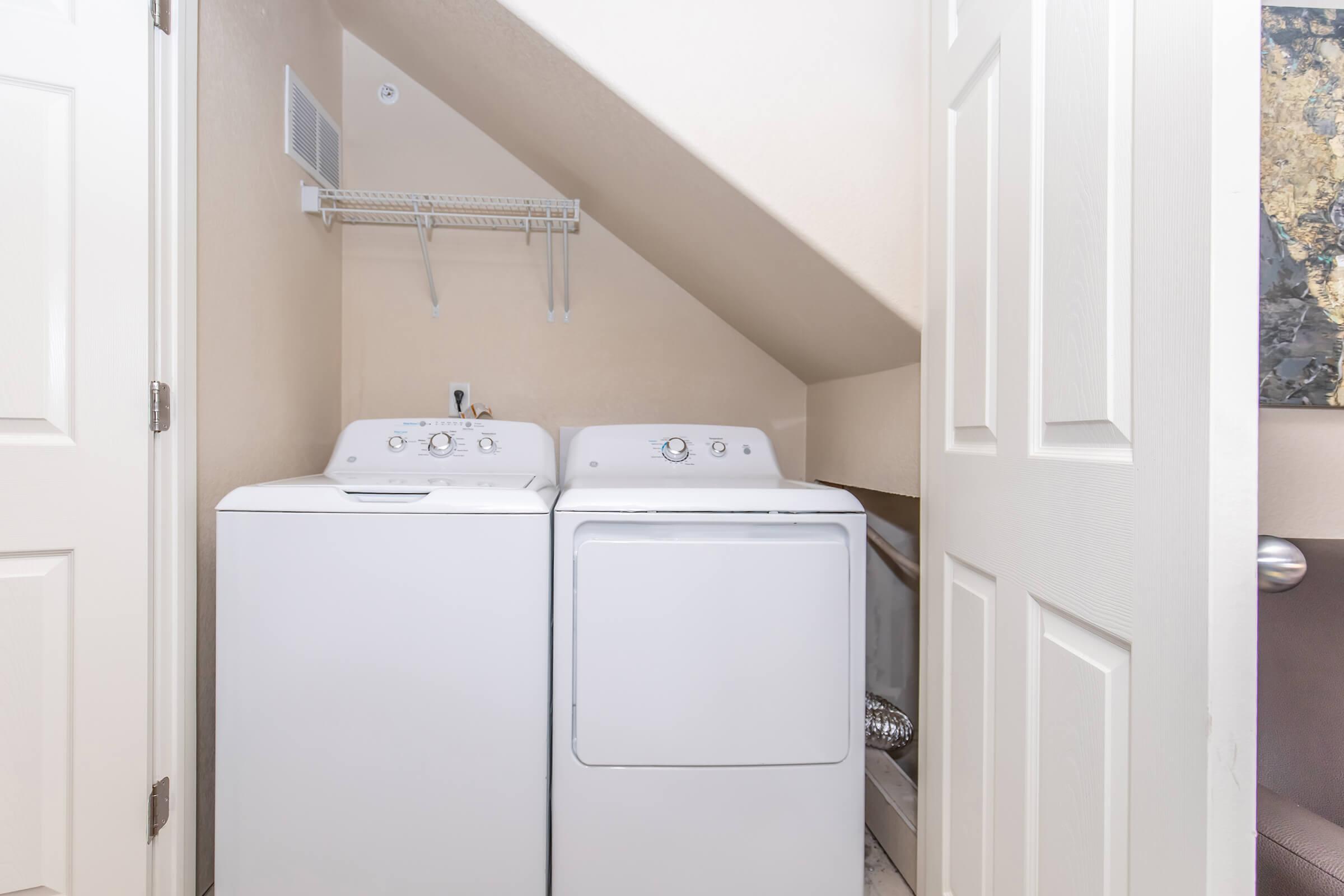 A small laundry area featuring a stacked washer and dryer in white. The space has light-colored walls and is located under a sloped ceiling. A wall-mounted shelf is above the appliances, and there is a close white door next to the laundry appliances.