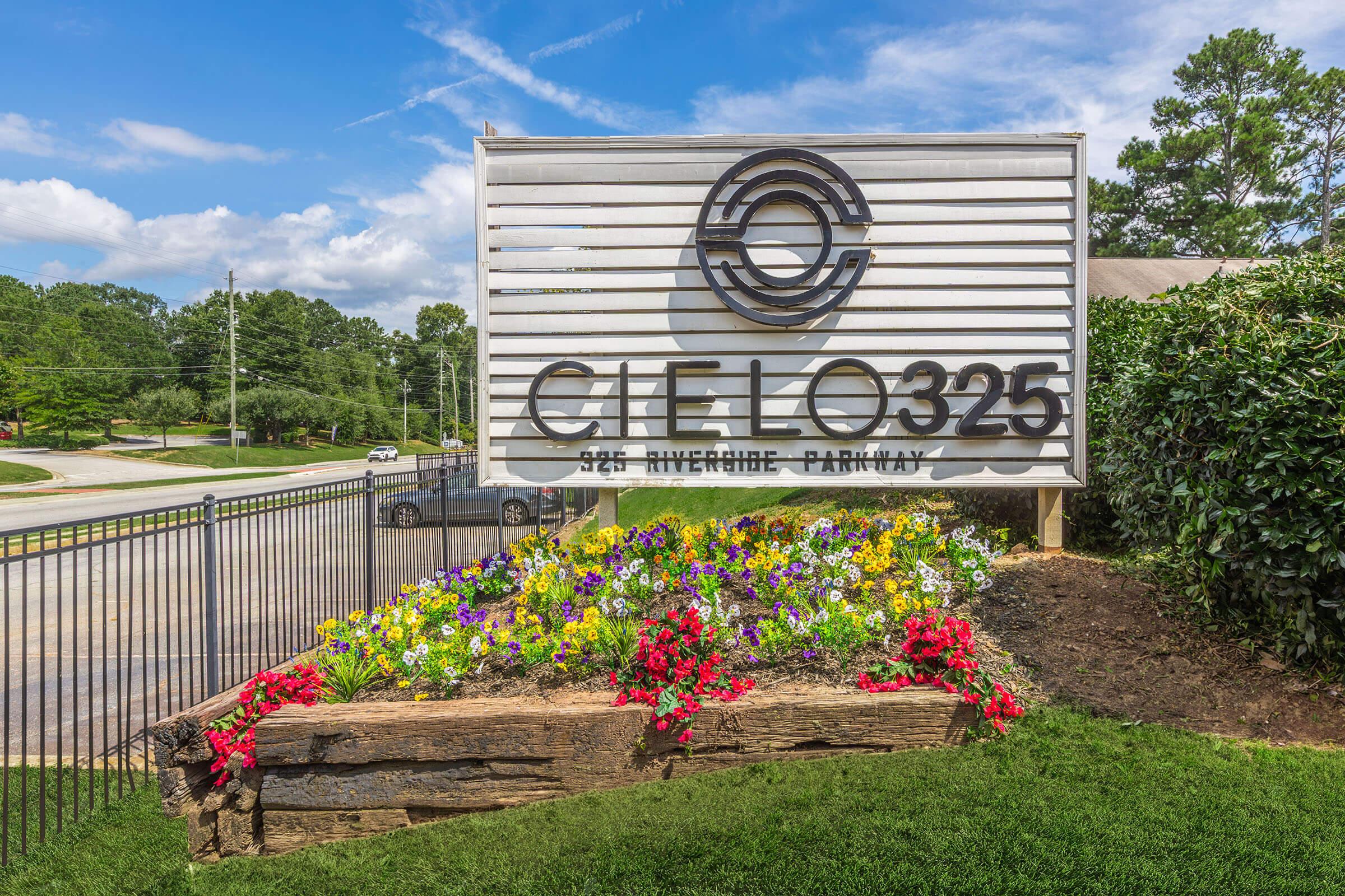 Sign for Cielo 325 located at 325 Riverside Parkway, featuring a circular logo at the top. The sign is surrounded by a flower bed with colorful blooms, set against a backdrop of green grass and trees under a blue sky with some clouds.