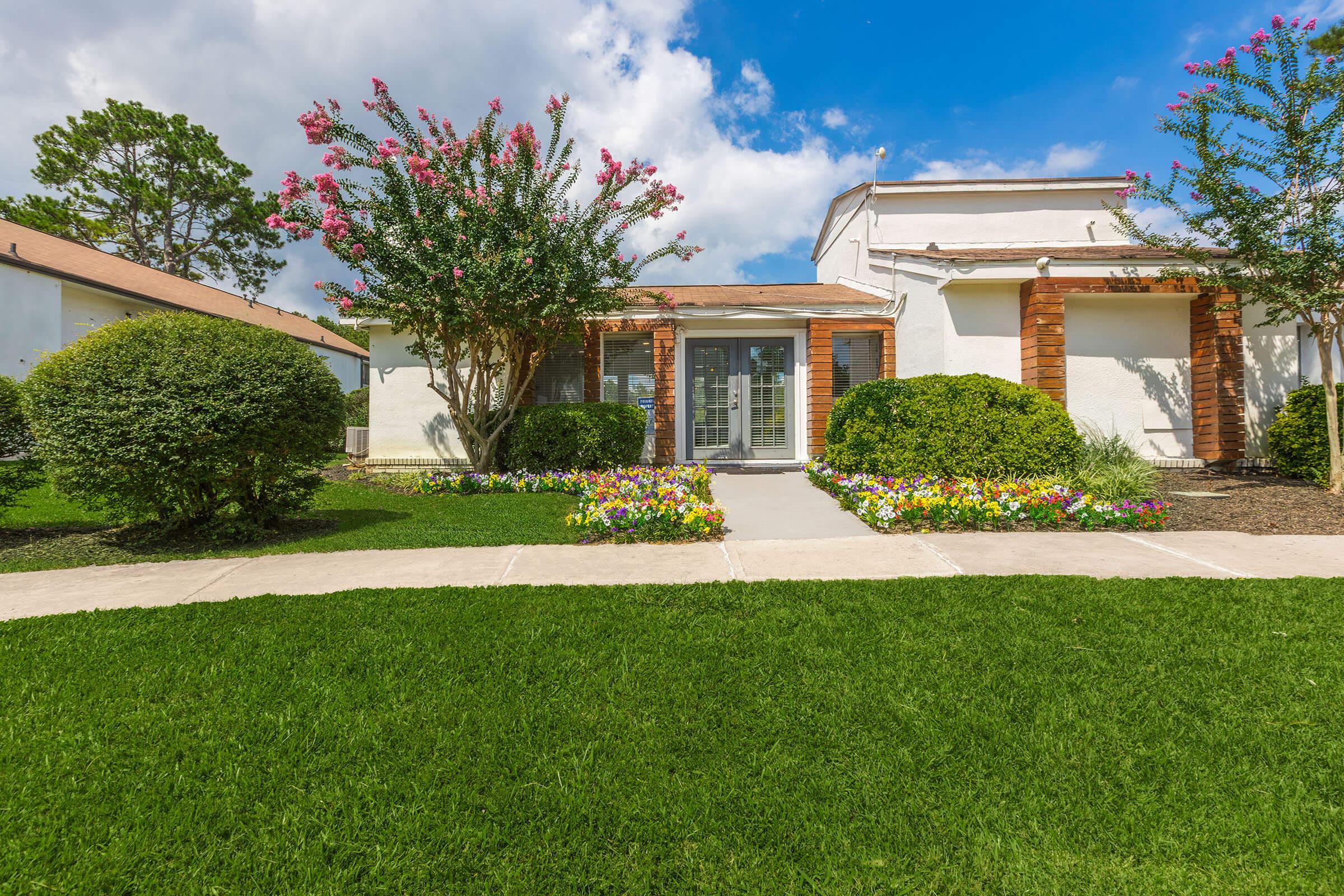 A well-maintained residential building with a lush green lawn and colorful flower beds in front. The entrance features a set of glass doors framed by decorative wooden accents, surrounded by neatly trimmed bushes and vibrant flowers under a blue sky with scattered clouds.
