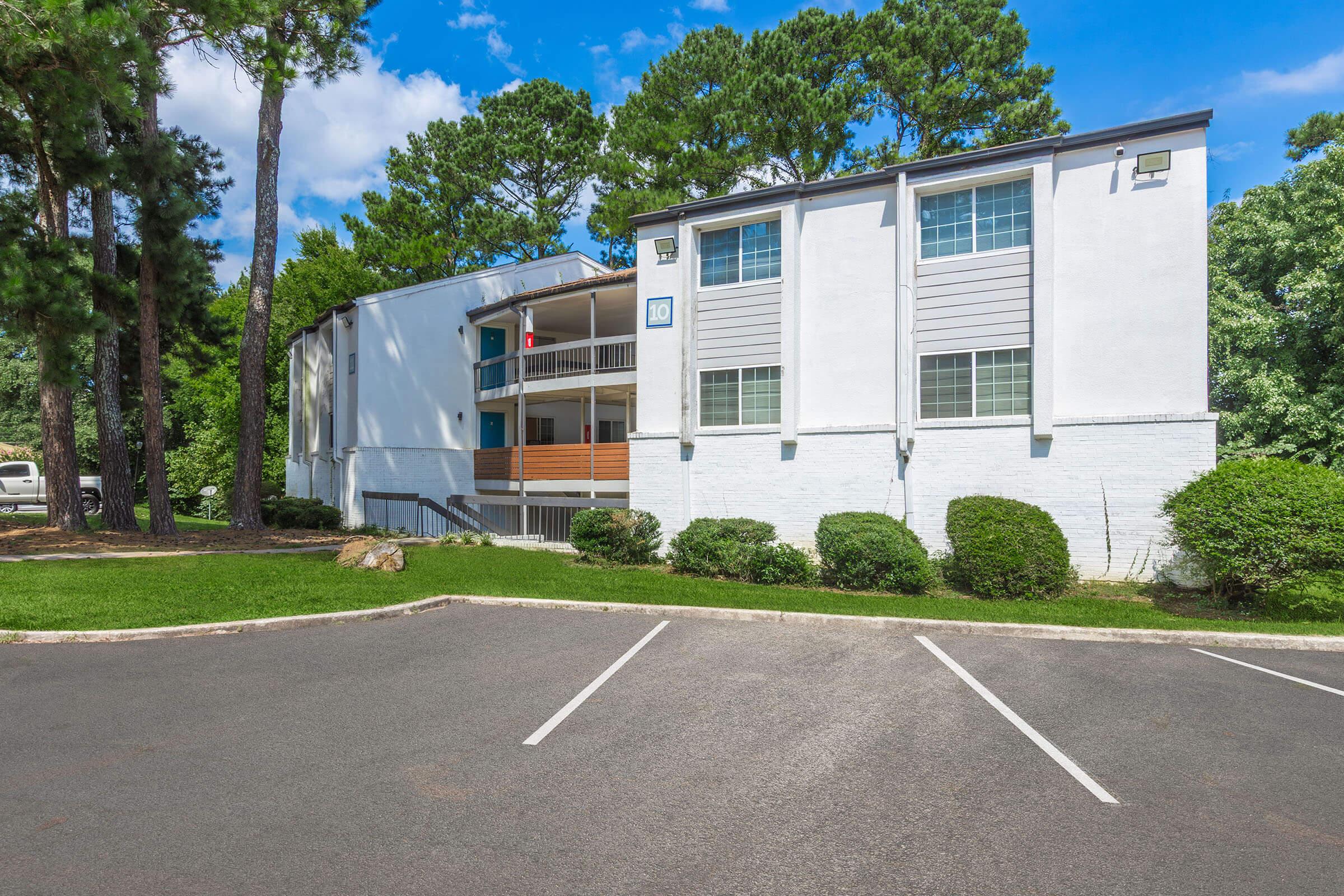 A modern two-story apartment building with a white exterior and gray accents, surrounded by lush green trees and shrubs. The building features balconies, parking spaces in the foreground, and a clear blue sky with a few clouds above.
