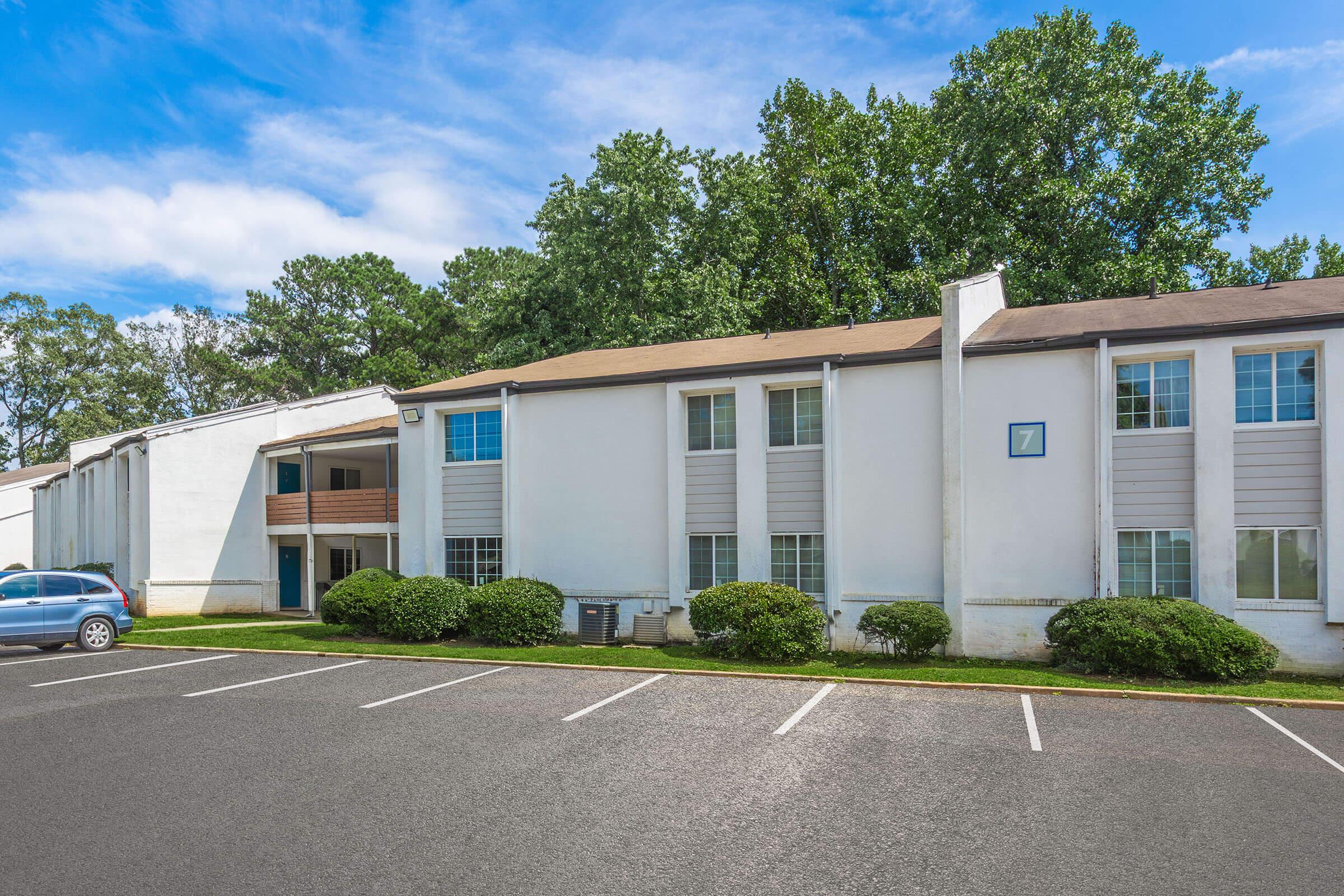 A two-story, white building with multiple windows and a number 7 visible on the side. The building is surrounded by green shrubs and a parking lot with empty spaces. Trees are visible in the background under a partly cloudy sky.