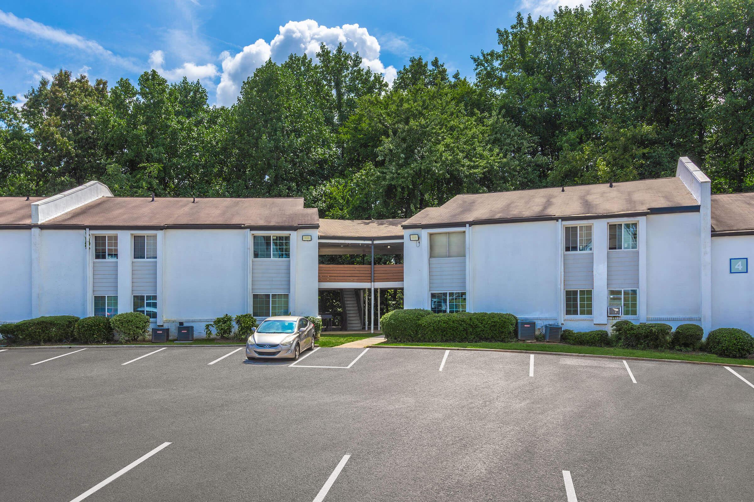 A two-story apartment complex with white exterior walls surrounded by greenery. The building has multiple windows and a central entrance. In the foreground, there is a parked silver car in an empty parking lot with marked spaces. The sky is clear with a few clouds.