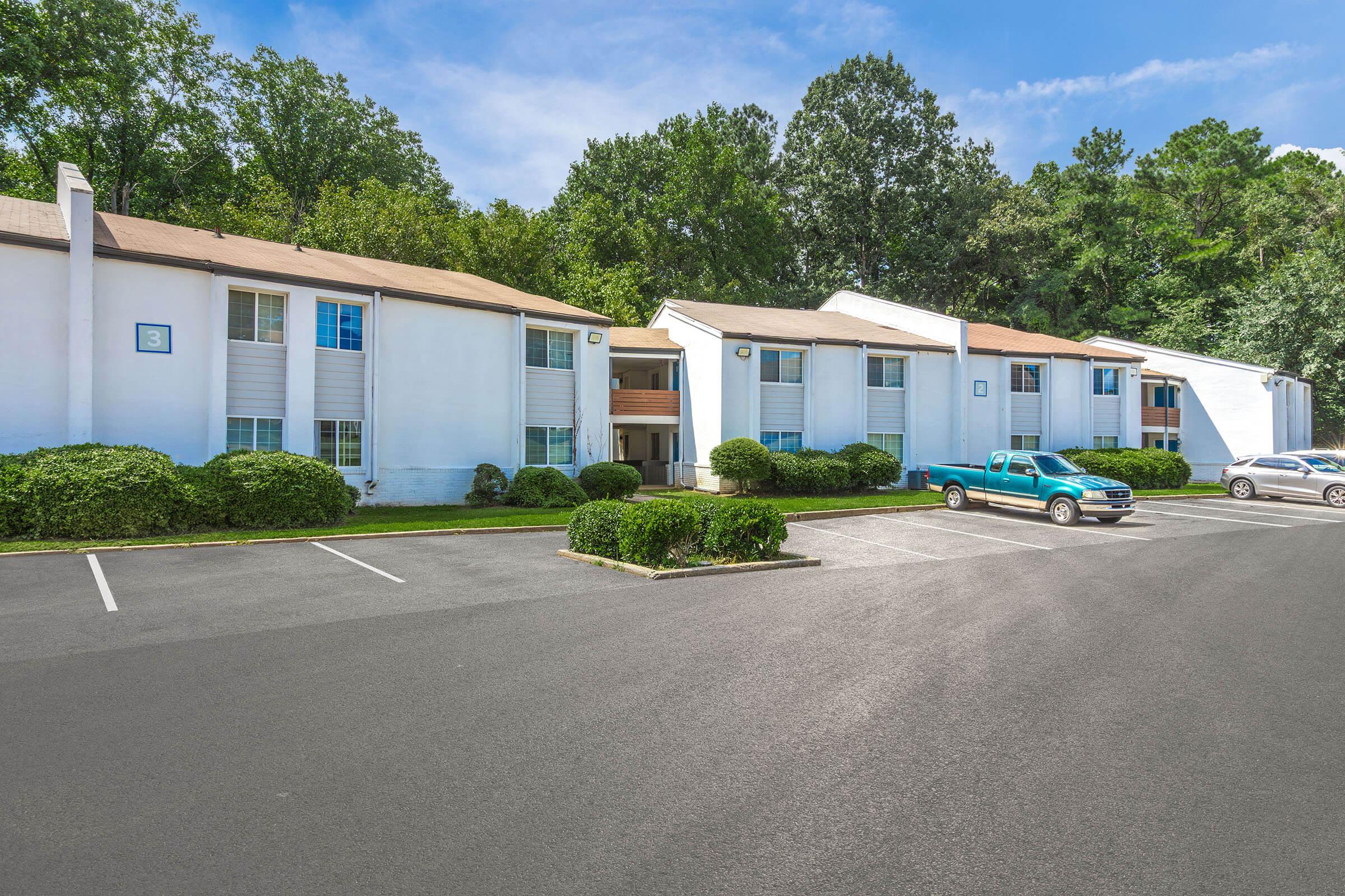 A row of white two-story apartment buildings surrounded by well-maintained greenery. The parking lot in front features several cars, including a blue pickup truck. Tall trees and a clear blue sky provide a pleasant backdrop.
