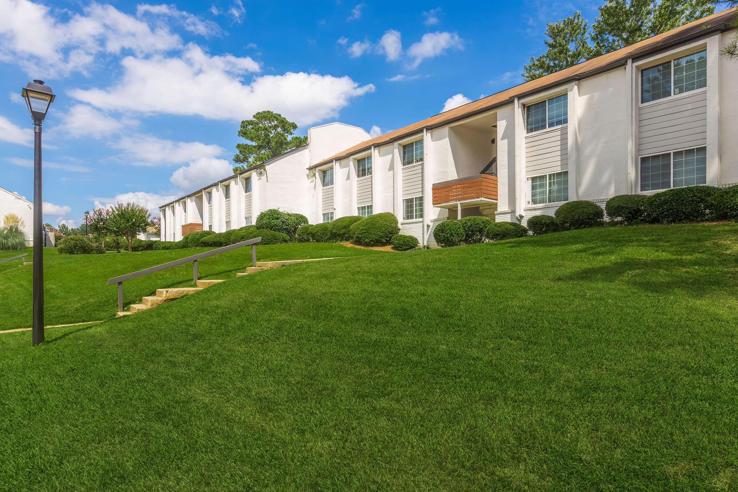 A well-maintained apartment complex with white exterior walls and large windows, surrounded by lush green grass and shrubs. A sloped lawn leads up to the building, with a pathway and lamppost in the foreground. The sky is bright blue with scattered clouds.