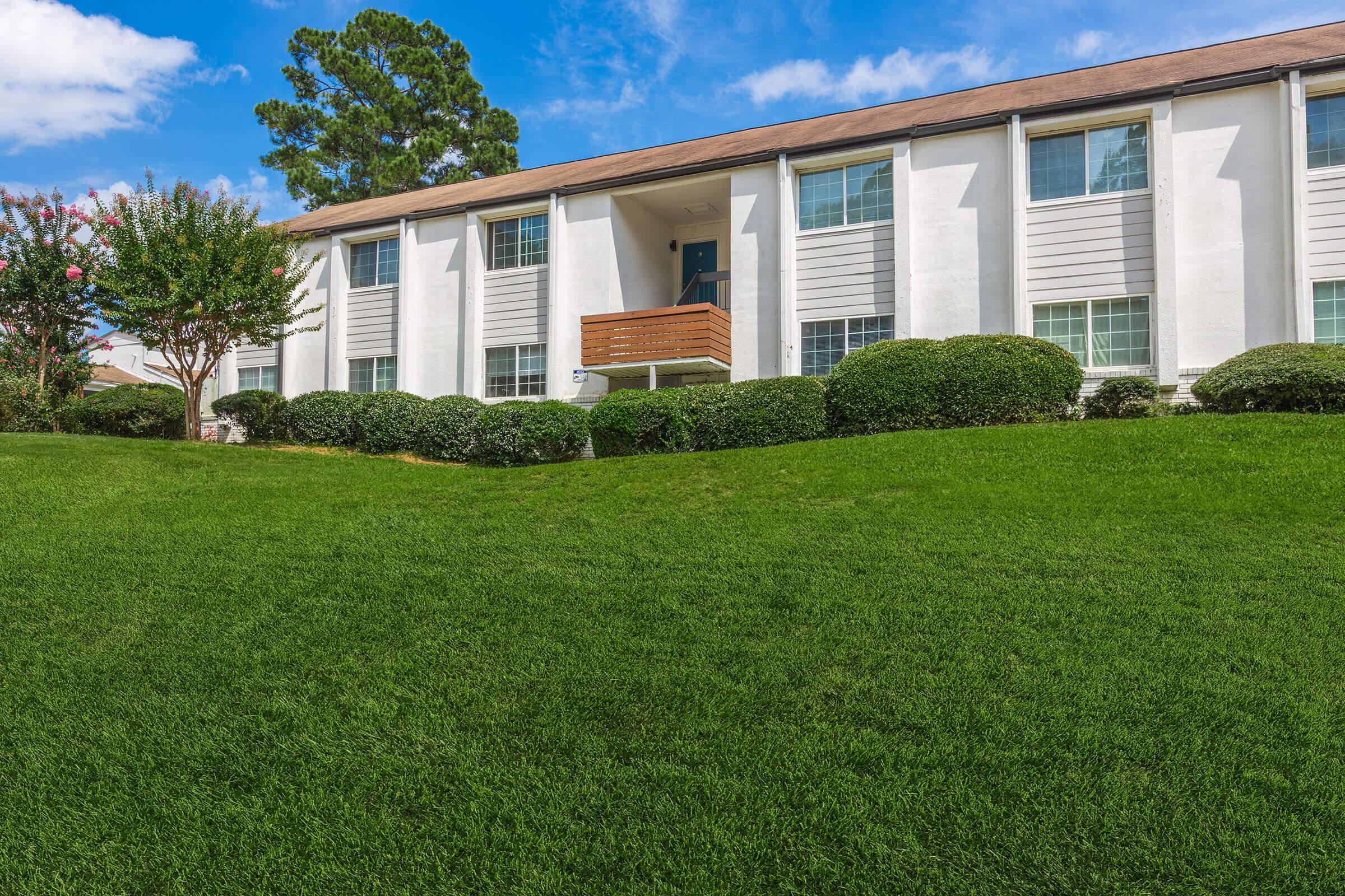 A well-maintained two-story apartment building with white exterior walls and multiple windows. The building features a wooden balcony on the second floor, surrounded by manicured green lawns and shrubs. Blue sky with a few clouds is visible in the background.