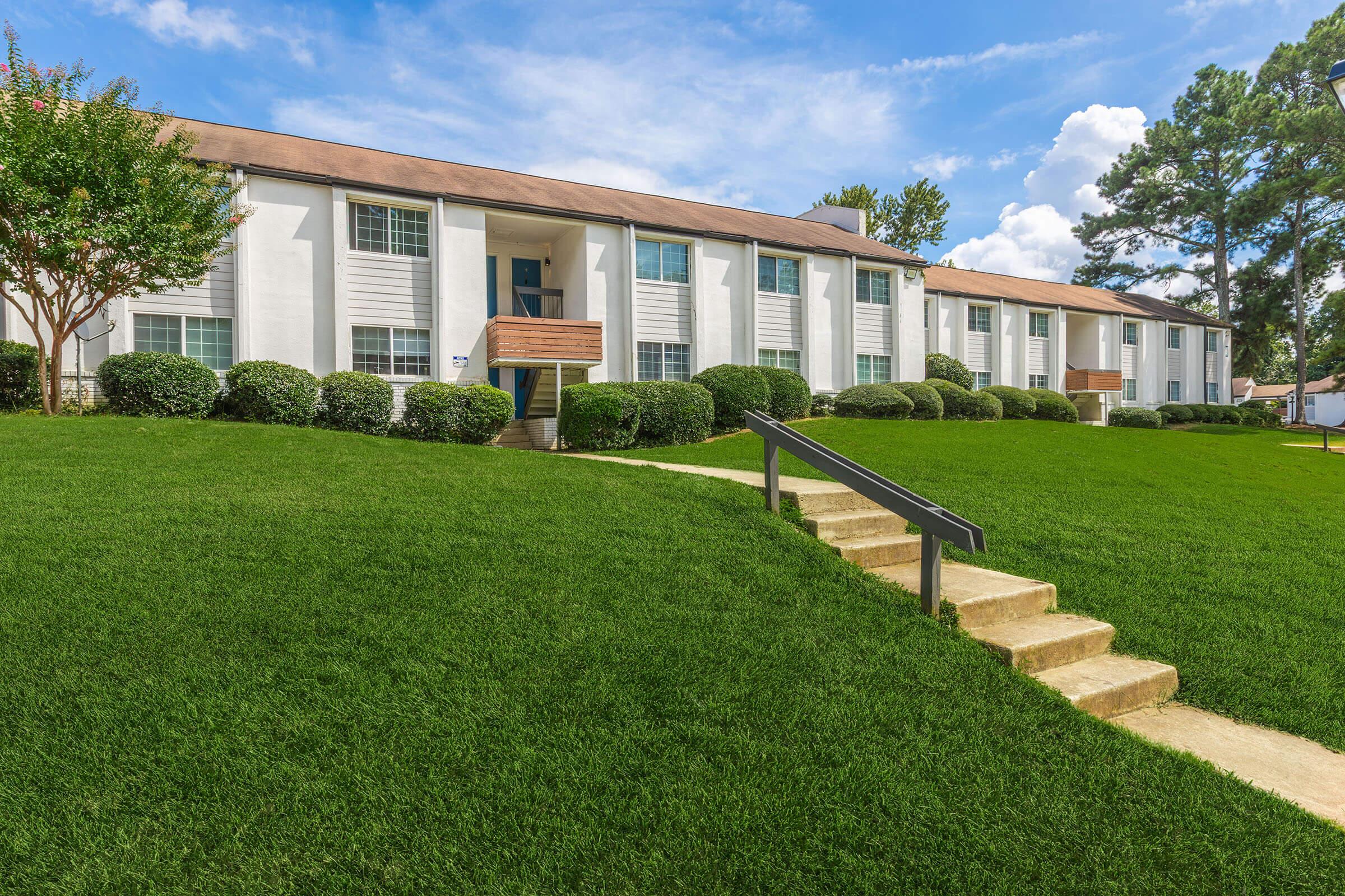 A well-maintained apartment building with multiple white units, surrounded by manicured green lawns and shrubs. There are concrete steps leading to the entrance, and the sky is blue with a few clouds, indicating a pleasant day.