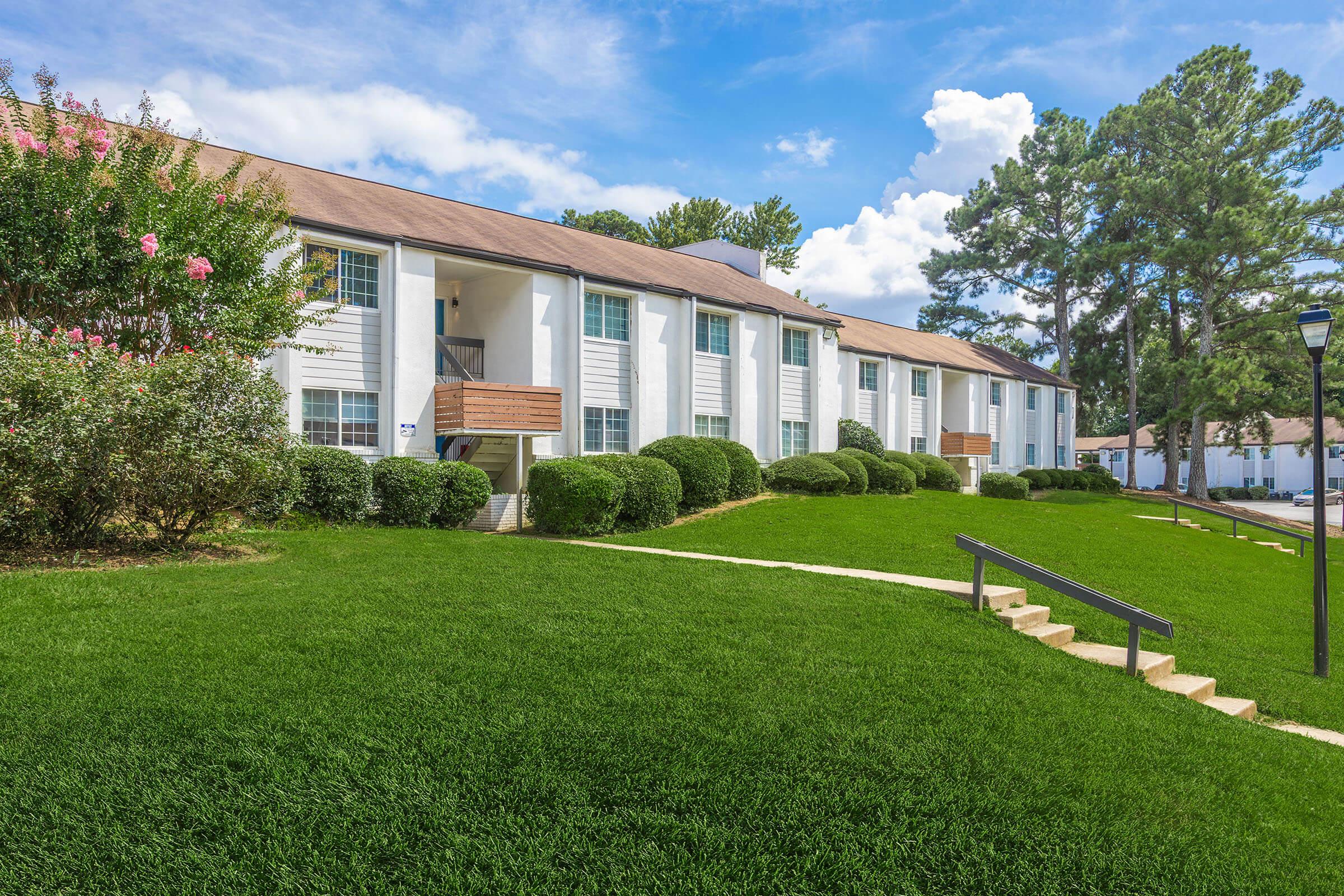 A well-maintained apartment complex with white exteriors, featuring multiple units and brown balconies. The landscaping includes manicured green lawns and shrubs, with a pathway leading to the entrance. Trees are visible in the background under a bright blue sky with fluffy clouds.