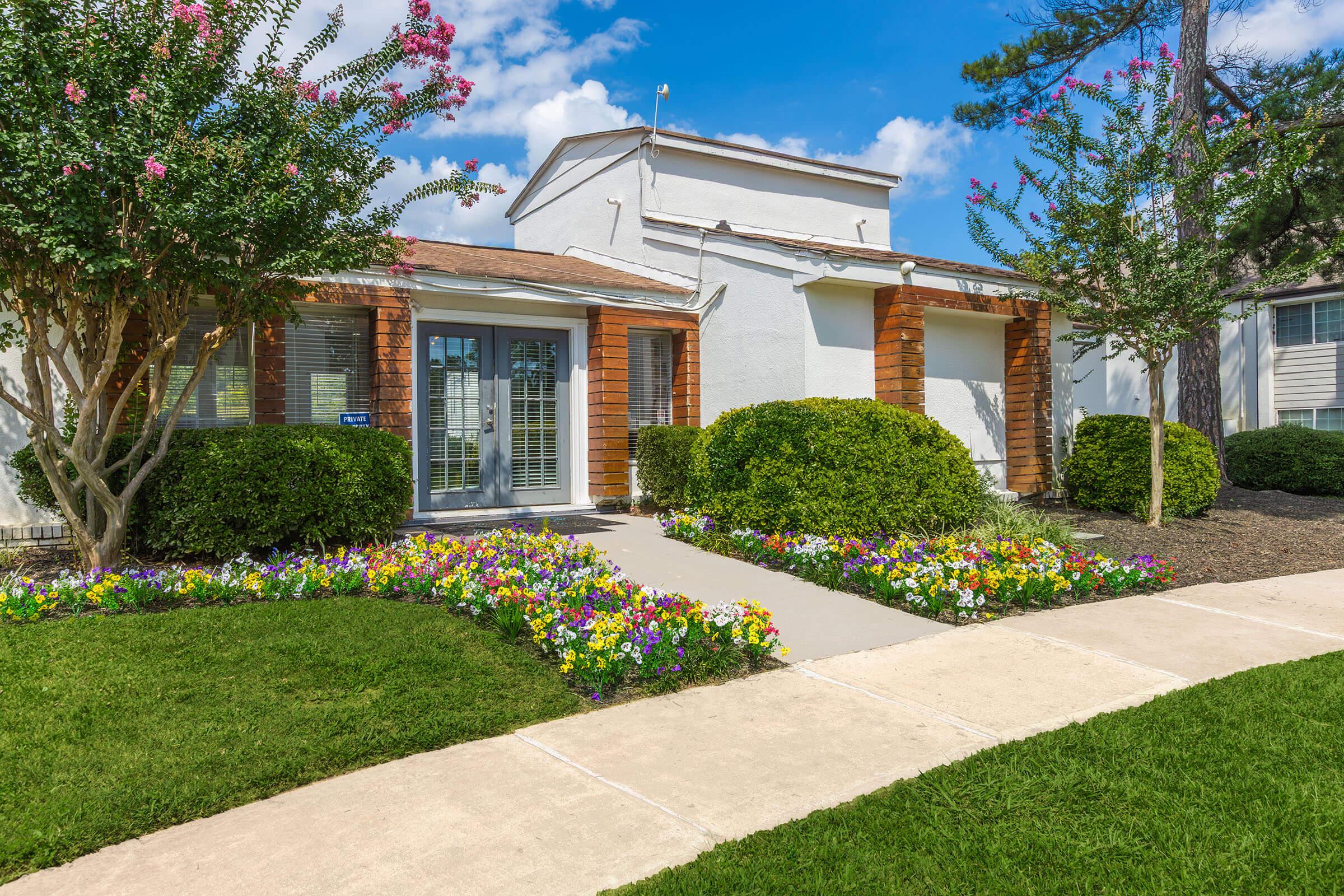 A well-maintained residential building with a landscaped front yard featuring colorful flower beds. The entrance is framed by two brick columns, with green shrubs and grass surrounding the path leading to the door. Bright blue sky and fluffy clouds complete the picturesque scene.