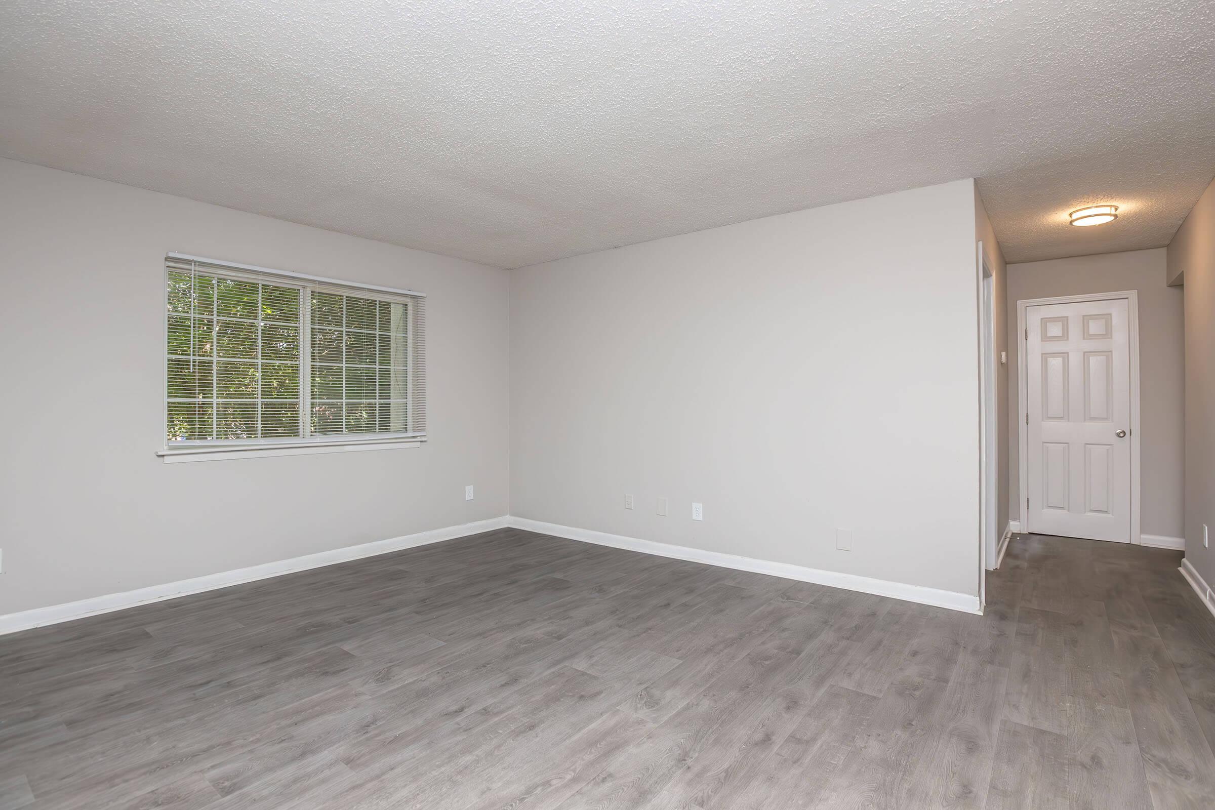 A vacant room with gray walls and a light-colored floor. It features a window with blinds, letting in natural light. On one side, there is a white door that likely leads to another area. The room is unfurnished and spacious, giving an open and airy feel.