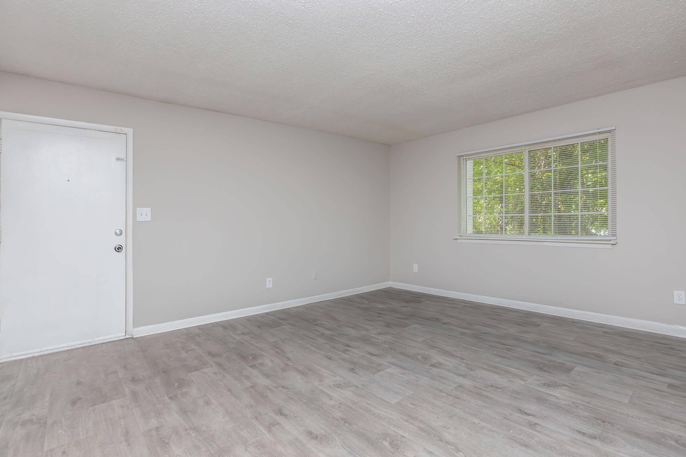 Empty room with light gray walls, a door on the left, and a window on the right allowing natural light. The floor features a light wood finish, creating a clean and spacious appearance.