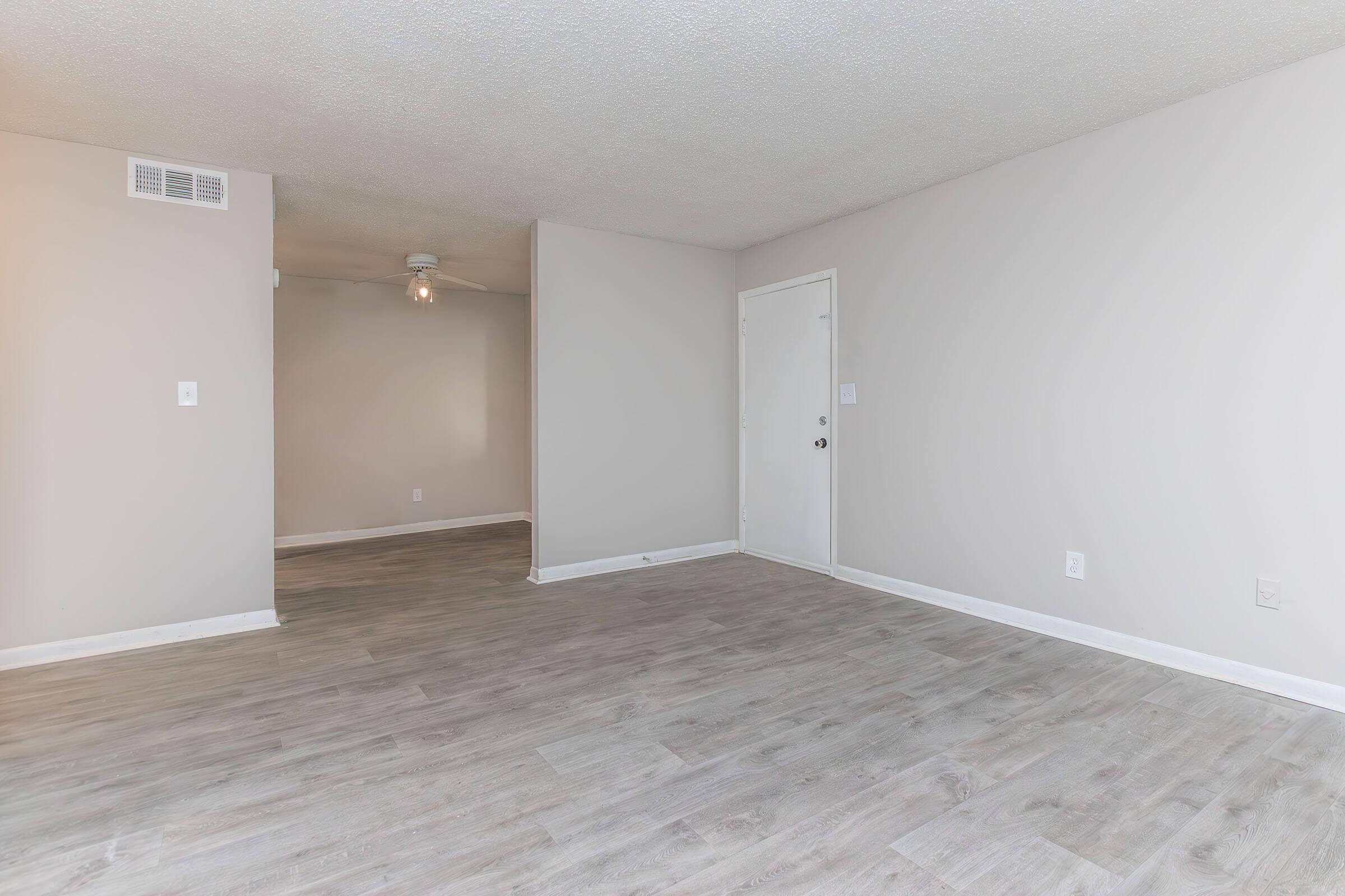 Empty room featuring light-colored walls, a ceiling fan, and a light-colored laminate floor. A door is visible on one side of the room, and another area is partially visible in the background. The space is well-lit and unfurnished, giving it a modern and open feel.