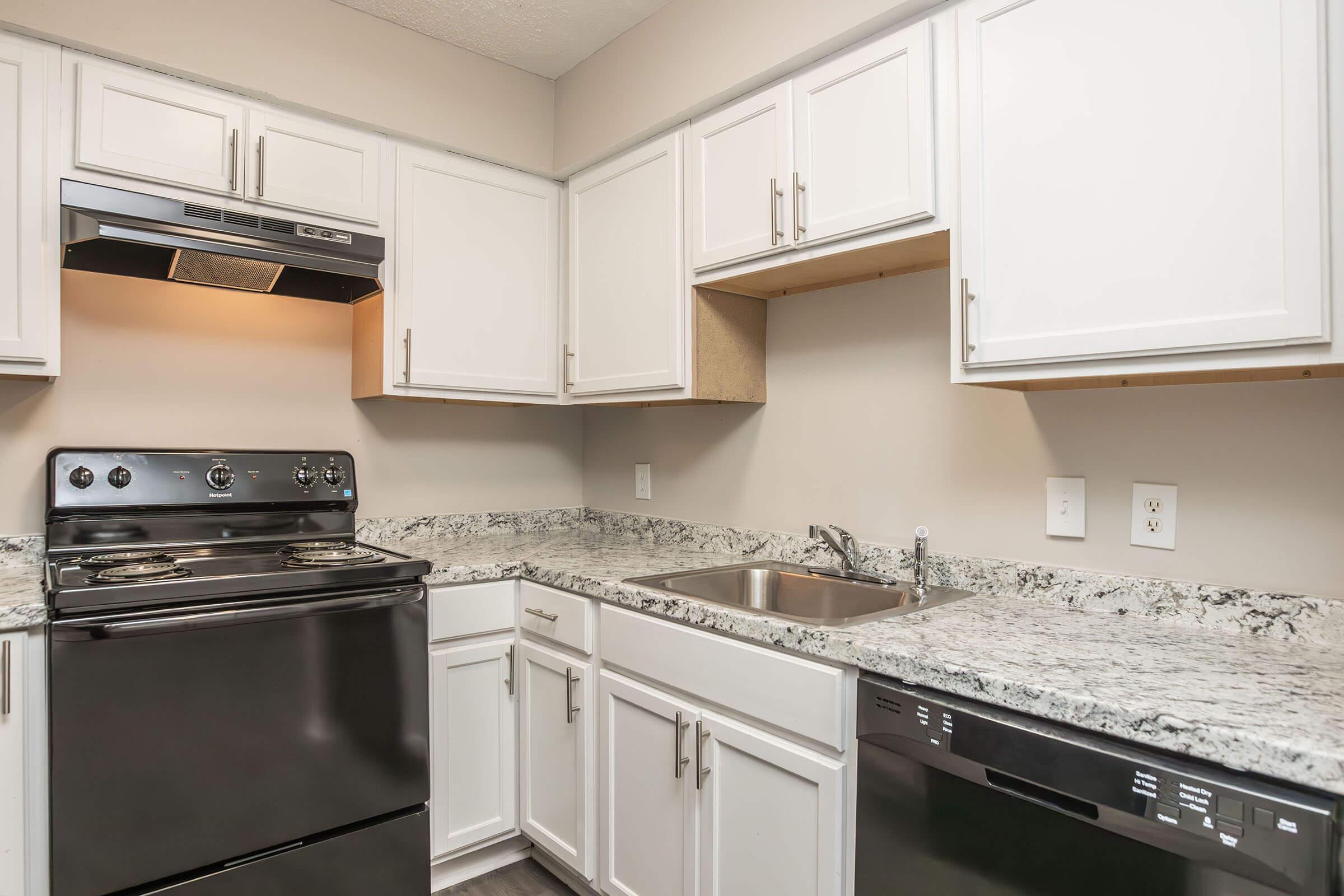A modern kitchen featuring white cabinetry, a granite countertop, a stainless steel sink, and black appliances, including a stove and dishwasher. The kitchen has neutral-colored walls and ample storage space, with a minimalist and clean design.